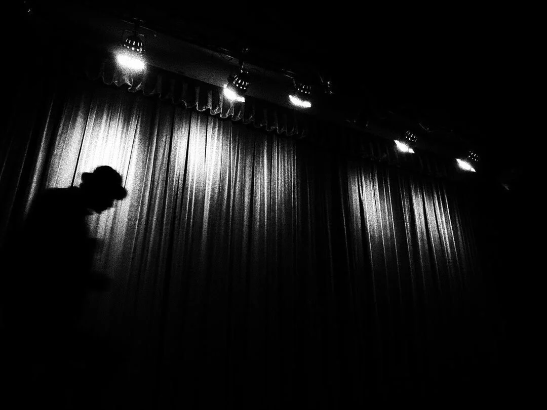 Silhouette of person in front of a lit stage curtain; black and white