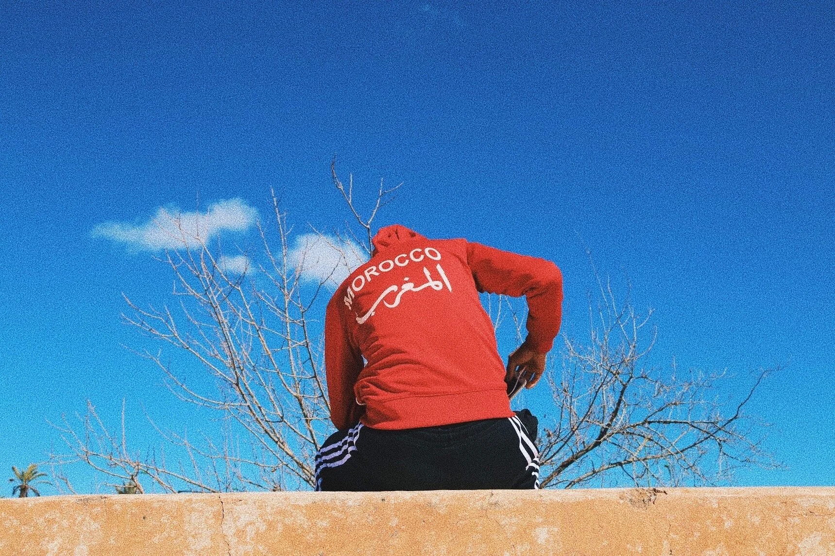 Person in red hoodie with 'Morocco' on back sitting on wall against blue sky.