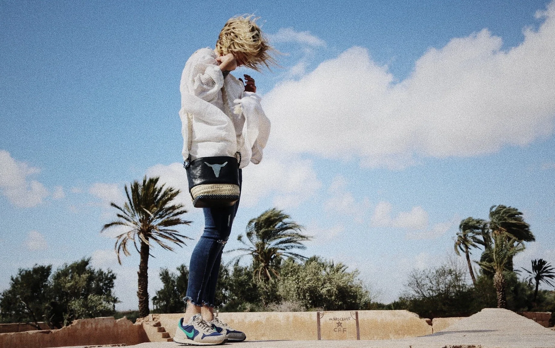 Person in jeans and white sweater on a rooftop with palm trees in the background