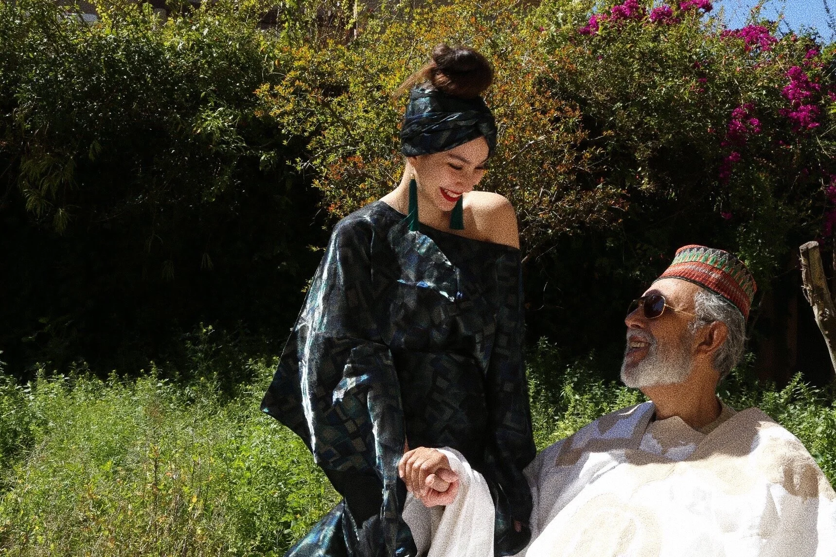 A woman and an older man in traditional attire holding hands outdoors in a garden setting.