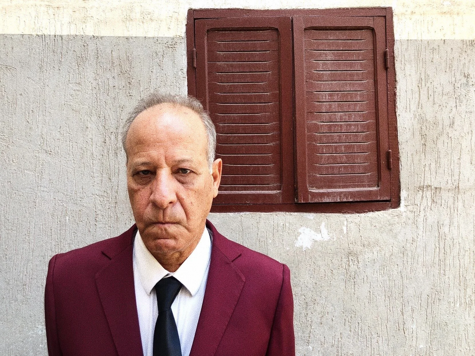 Man in a maroon suit with a black tie standing in front of a textured wall with a closed brown shuttered window.