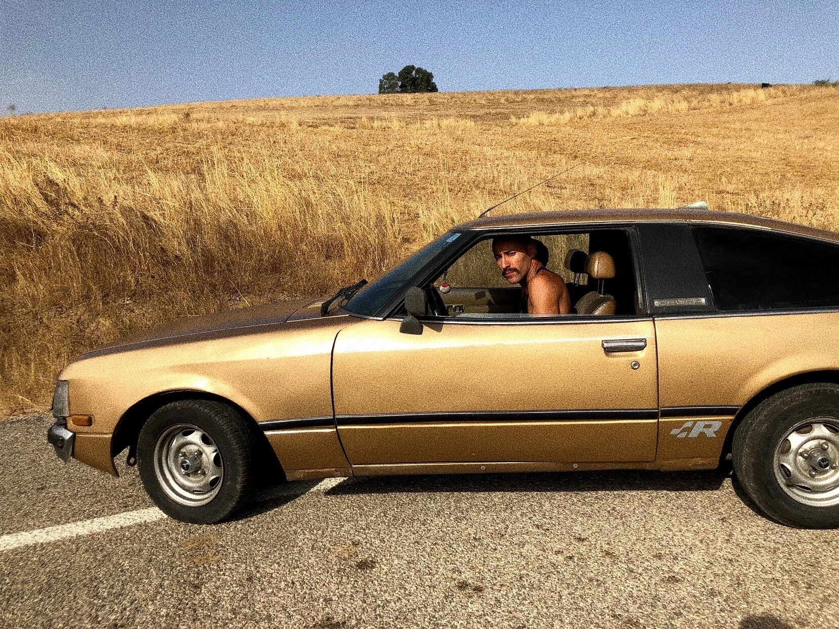 Vintage gold Toyota Celica car on a rural road with dry grass in the background, driver inside wearing a hat.