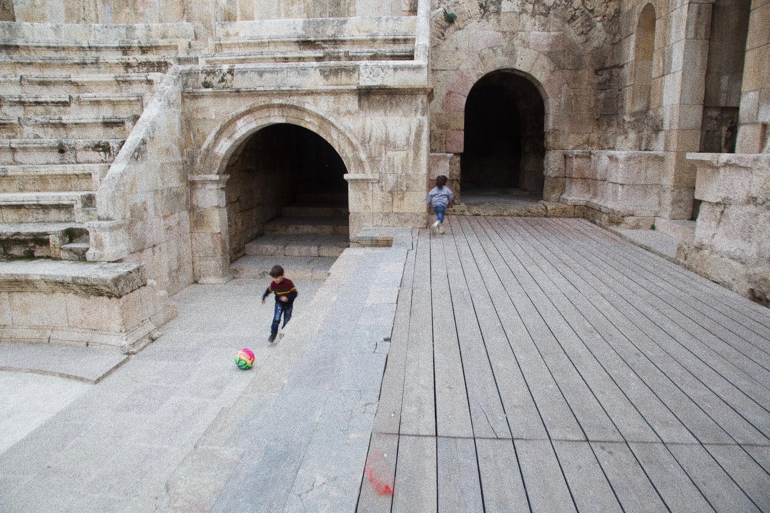 Two children playing near ancient stone arches, one boy kicks a colorful ball on a stone surface.