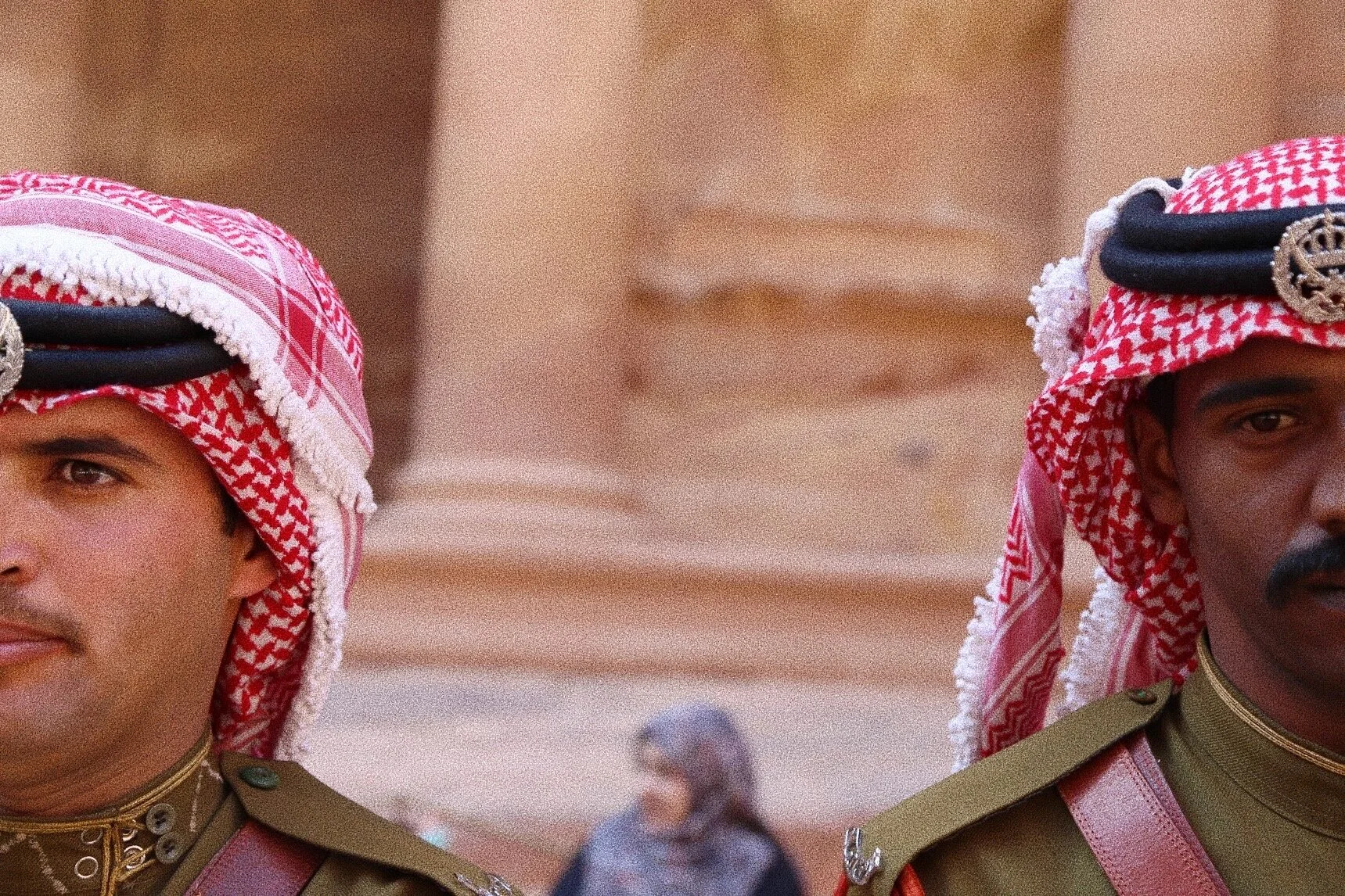 Two men in traditional Middle Eastern attire, wearing red and white keffiyehs, standing in front of ancient columns.