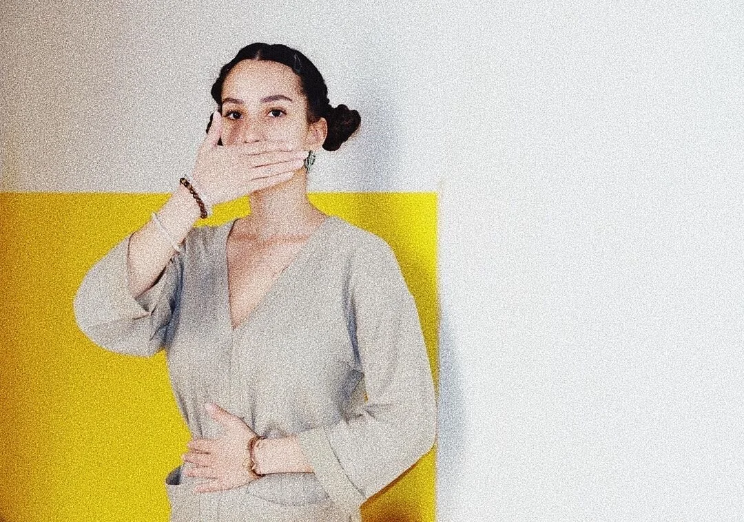 Person in gray outfit covering mouth with hand against a white and yellow background.