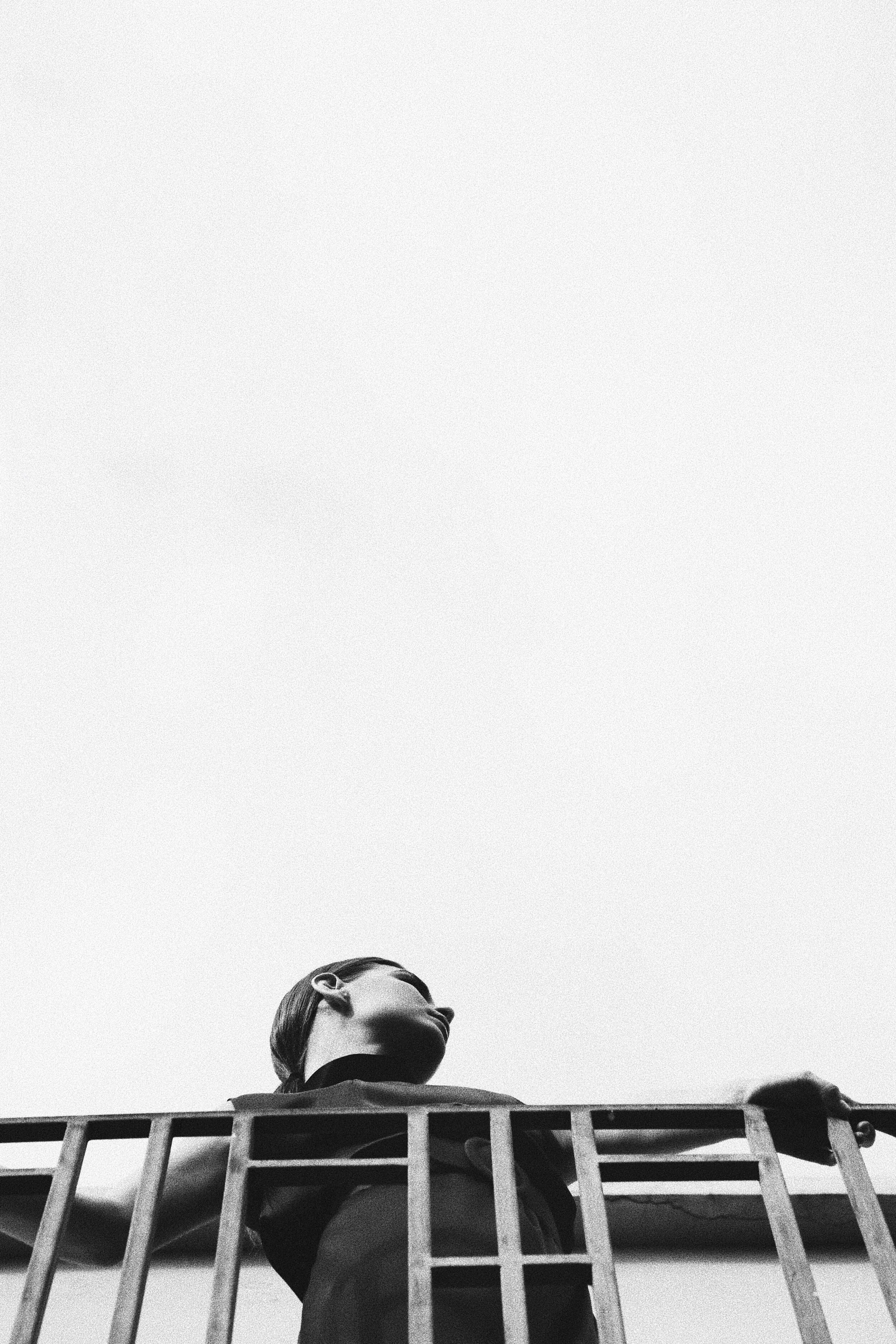 Black and white photo of a person leaning on a metal railing, viewed from below against a bright sky.