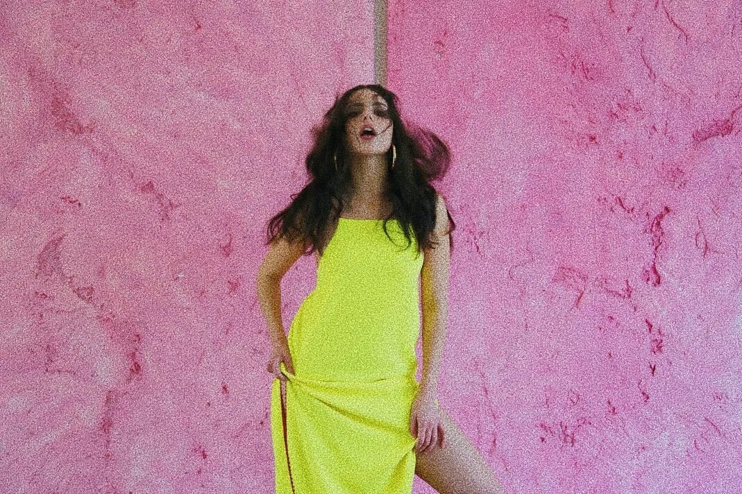 Person in a yellow dress standing against a pink textured wall.