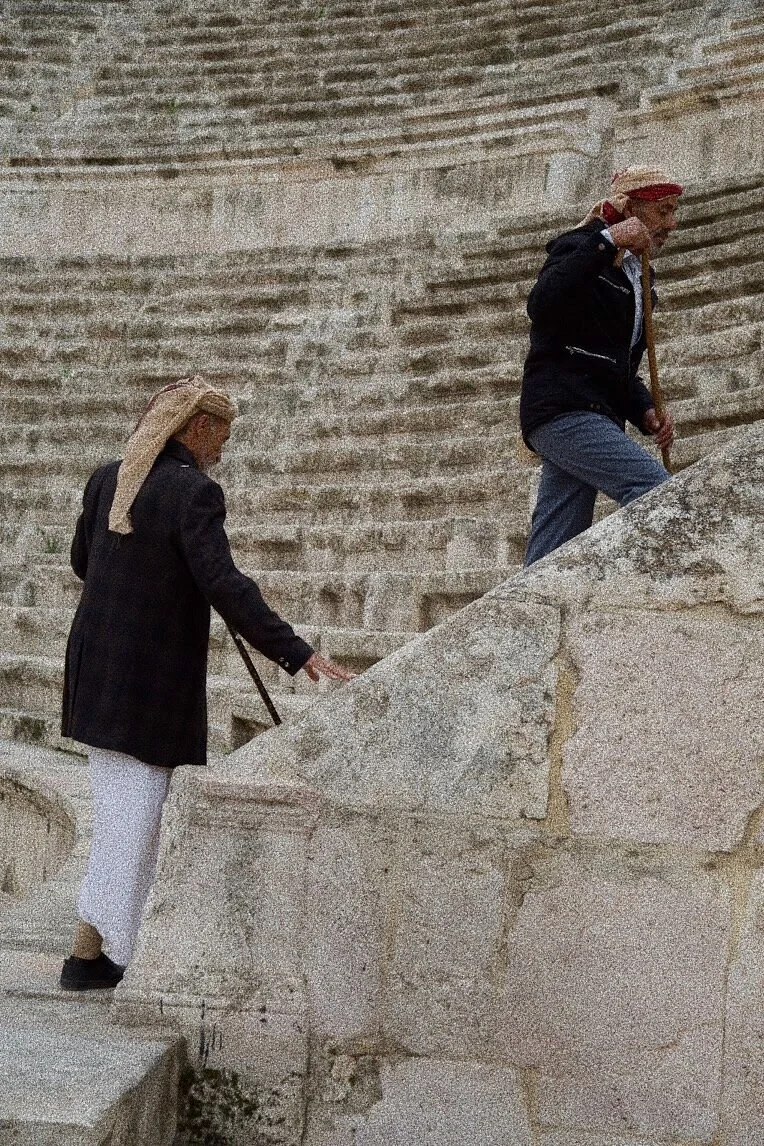 Two older individuals wearing traditional headwear, walking up stone steps in an ancient amphitheater.