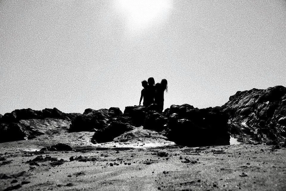 Silhouette of three people standing on a rocky beach with sunlight overhead in black and white.