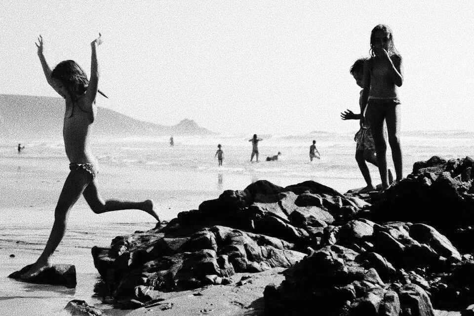Children playing on a rocky beach with others in the background by the shoreline.