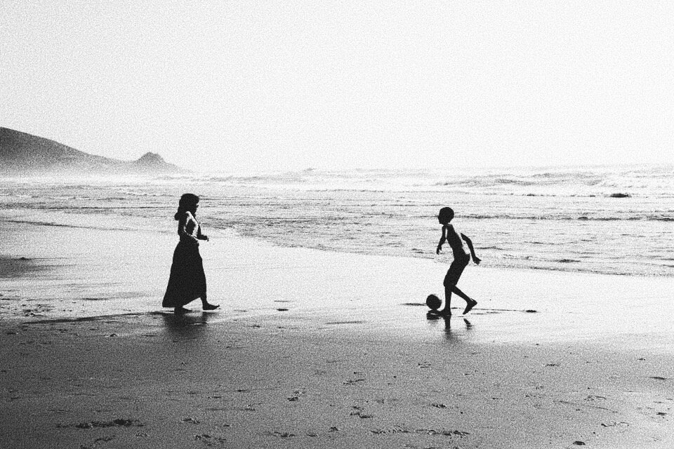 Silhouettes of two people on a beach, one kicking a soccer ball, with the ocean and hills in the background.