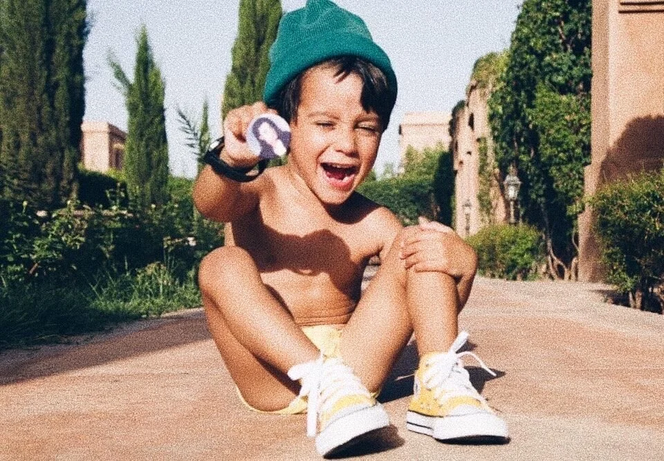 A young child wearing a green beanie, sitting on a path and laughing, while holding a small round object. The child is in a sunny outdoor setting with greenery and trees in the background, wearing white sneakers and shorts.