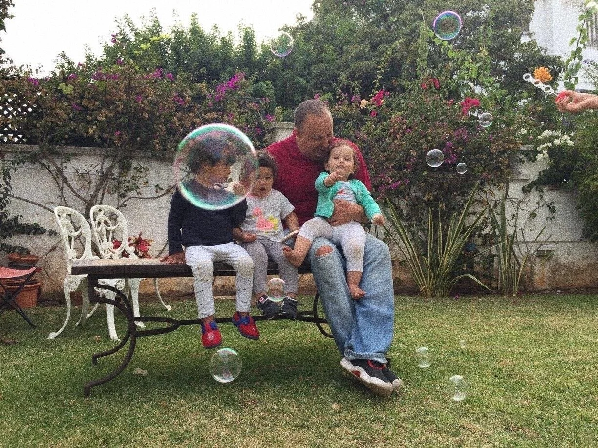 A family sitting on a bench in a garden with bubbles floating around. An adult is holding a child, and two more children are sitting nearby, looking at the bubbles. The garden has plants and a white lattice fence in the background.