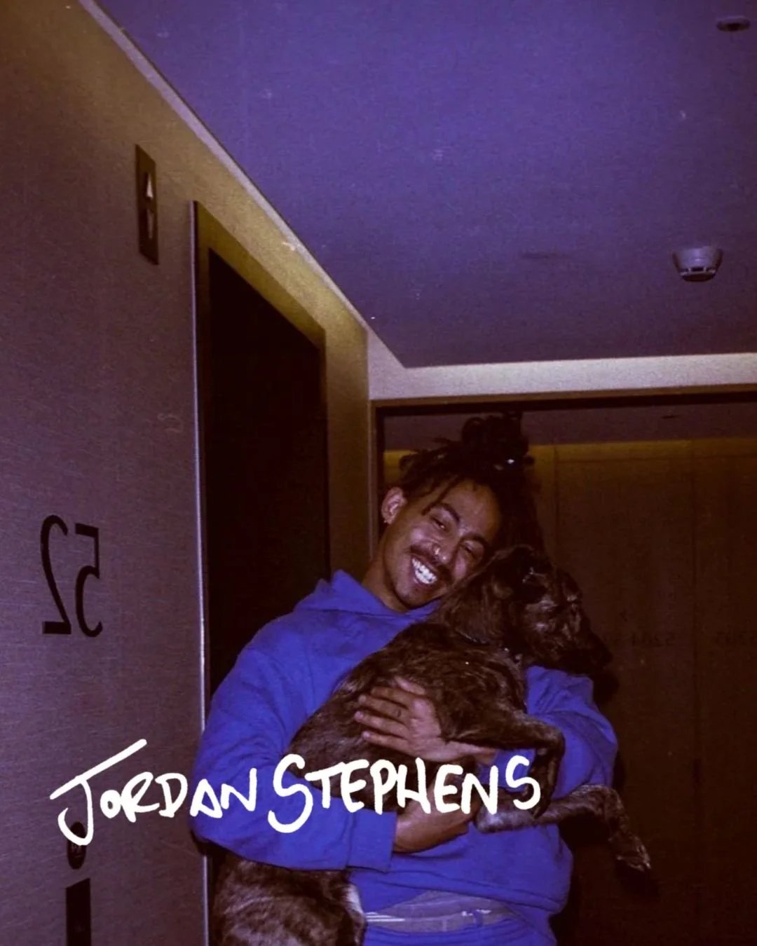 A person with dreadlocks and a septum piercing, smiling and hugging a dog.