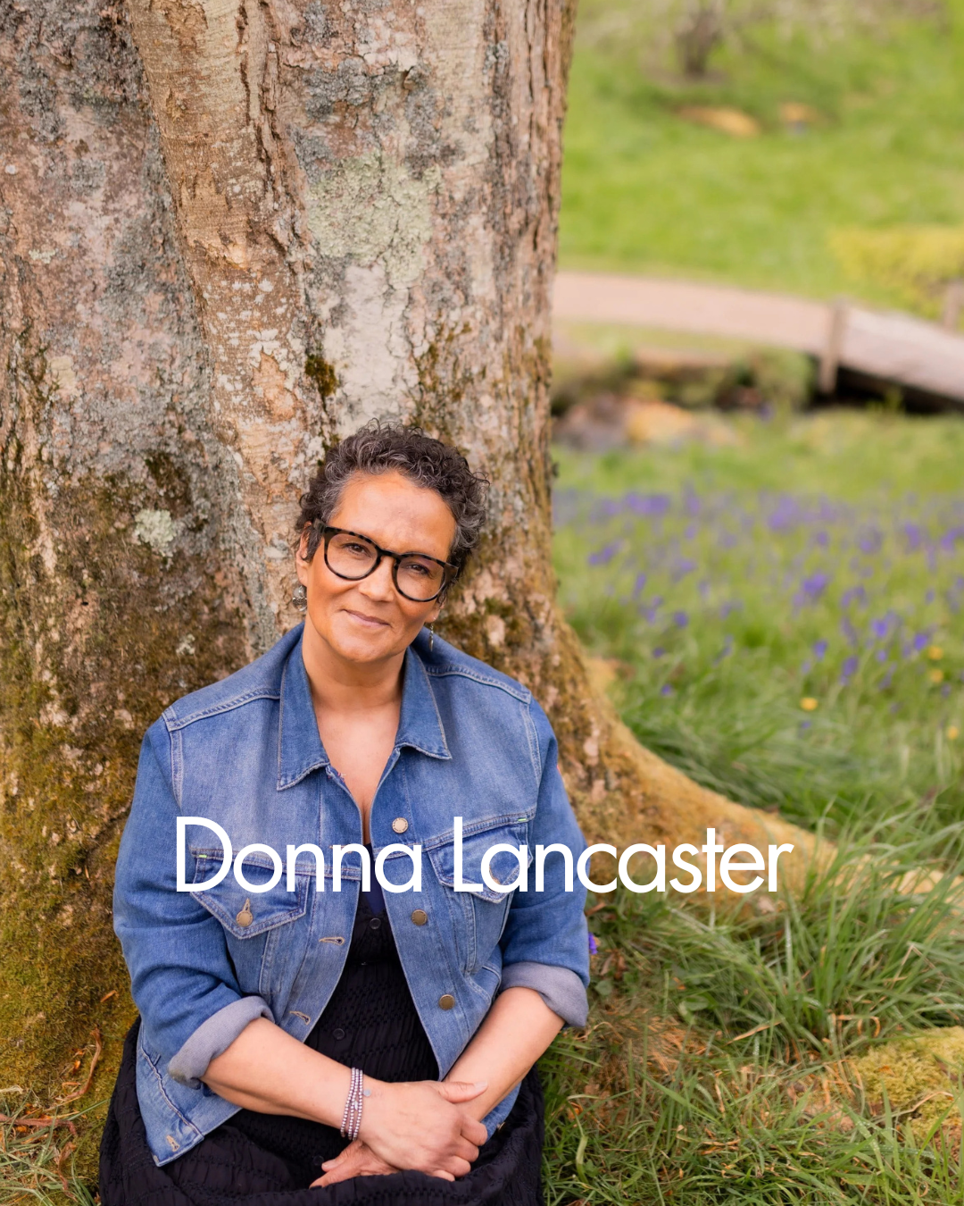 A woman with curly hair, glasses, and earrings, wearing a cozy sweater, standing outdoors behind a tree and smiling at the camera.
