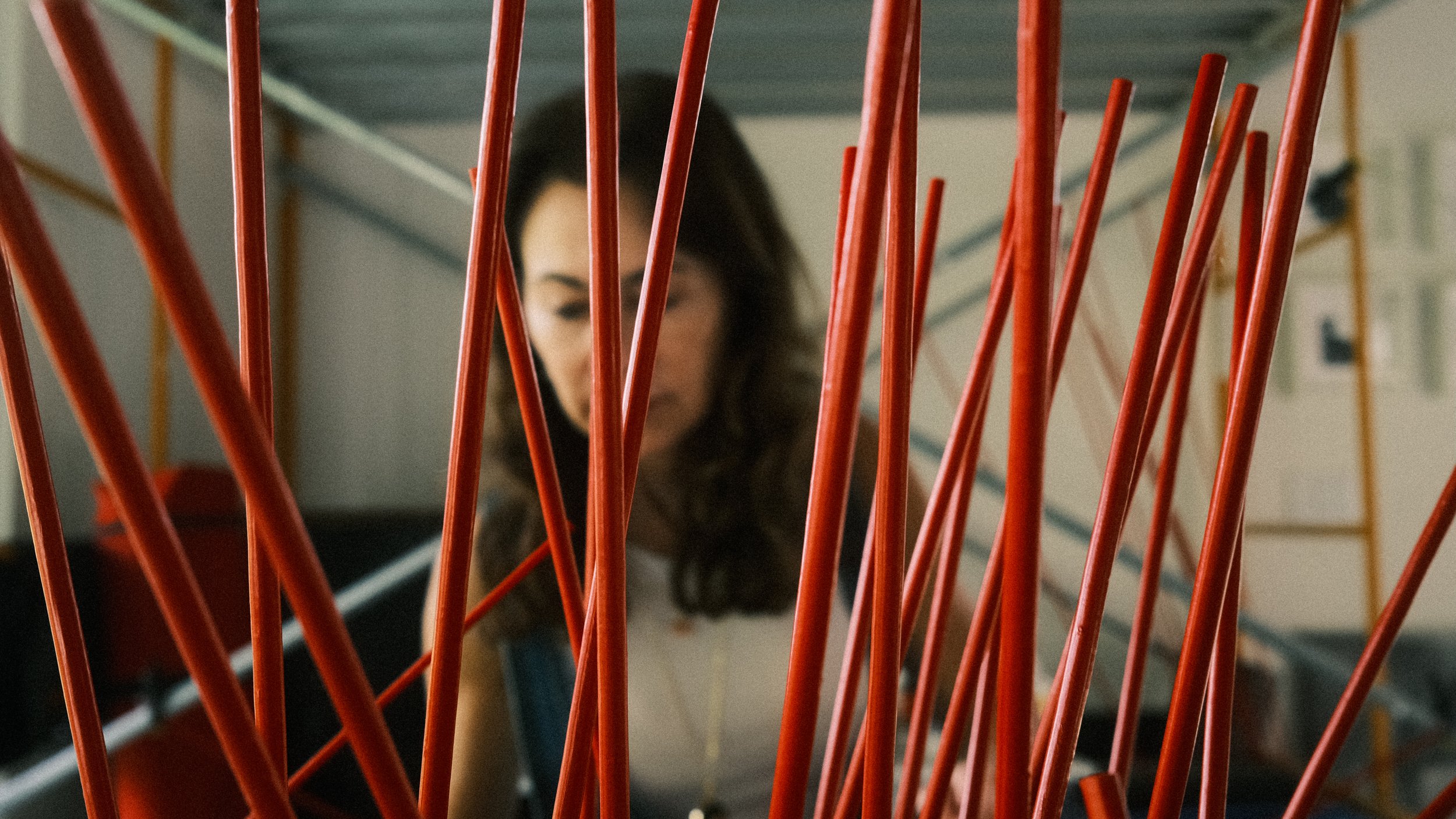 Detail of a red rod sculpture in progress, with Sylvia Riquezes working in the background.