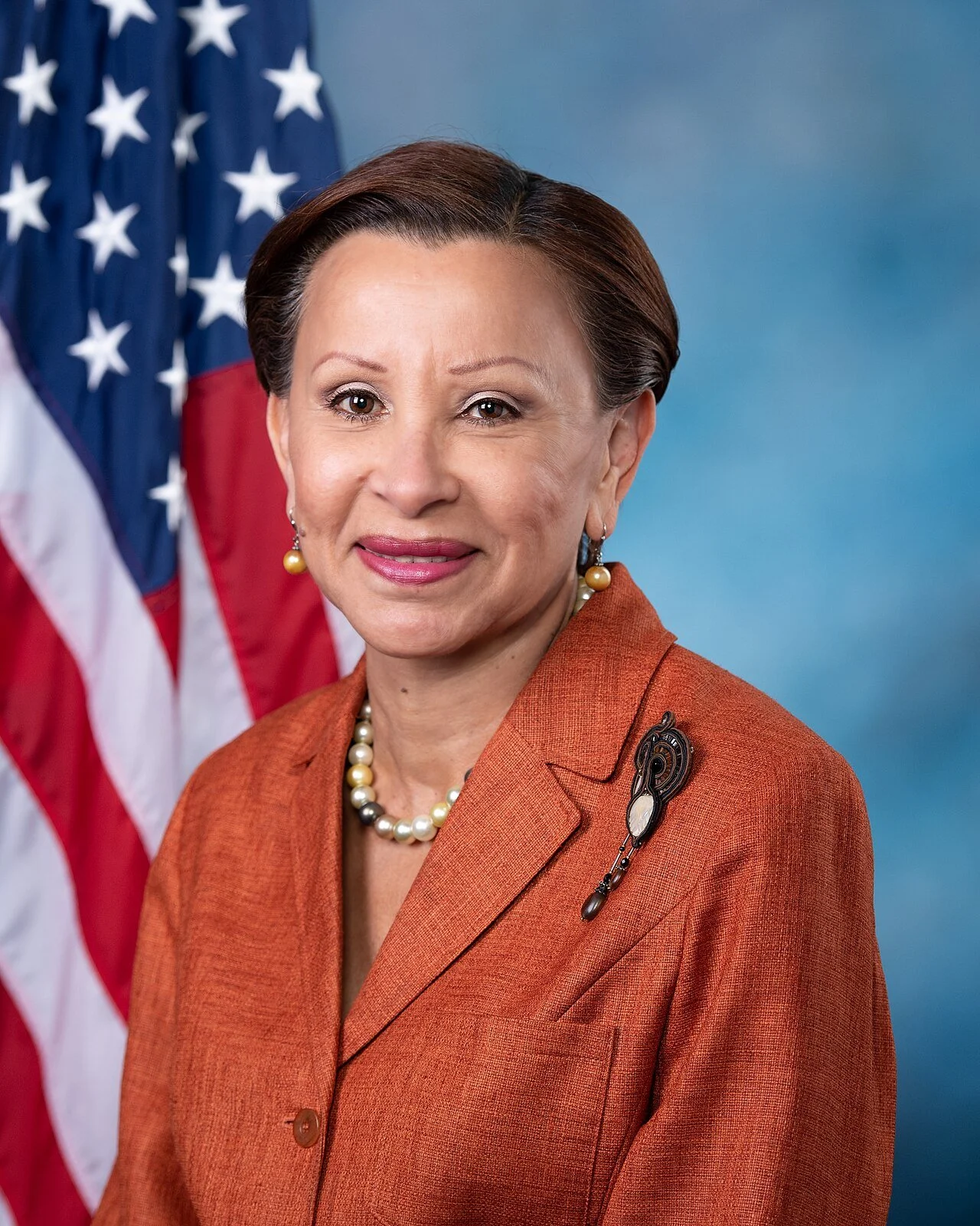 A woman with short brown hair wearing an orange blazer with a brooch, pearl necklace and earrings, standing in front of an American flag.