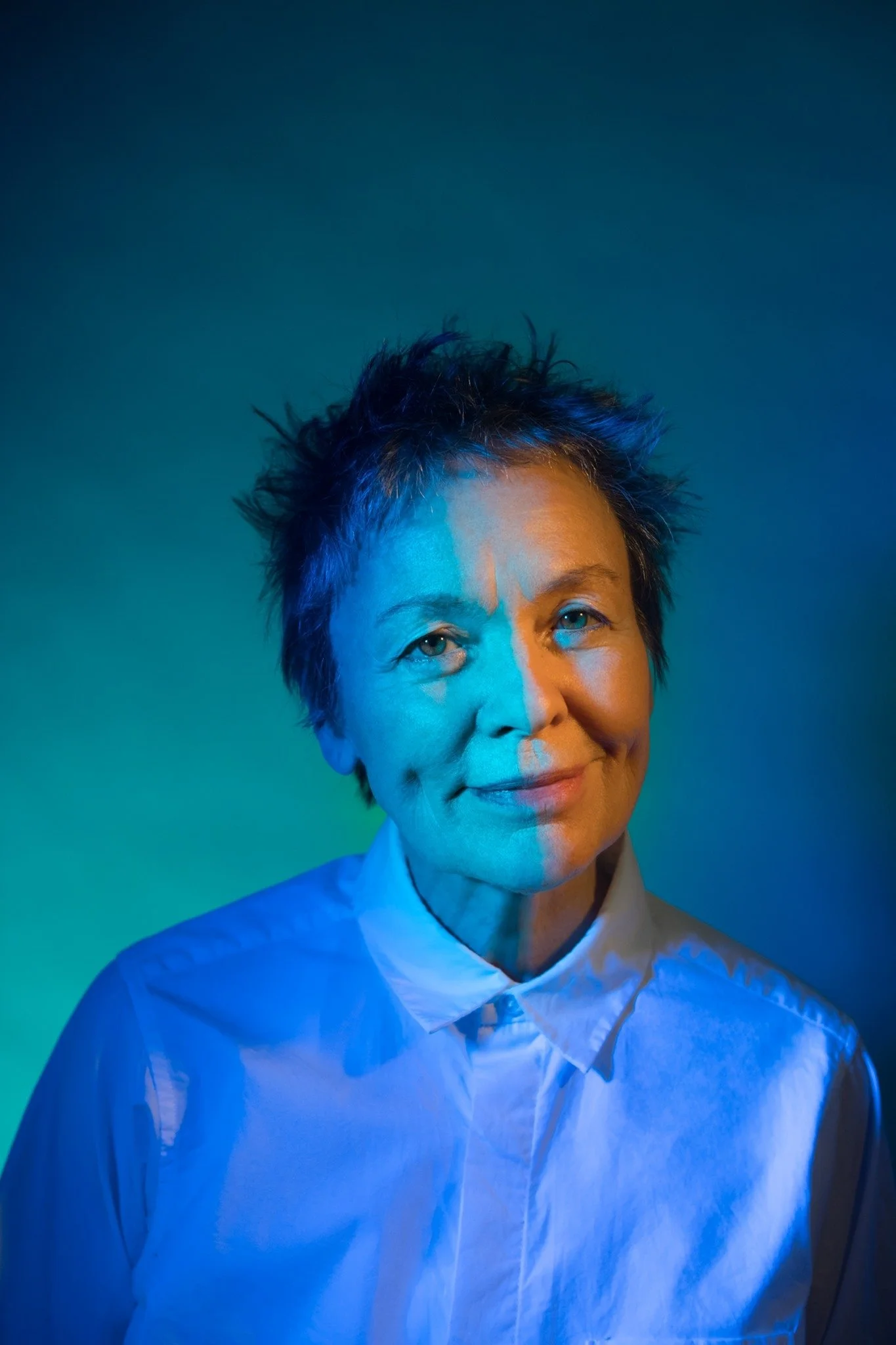 Portrait of a woman with short, spiky hair wearing a white shirt, illuminated by colorful blue and orange lighting.