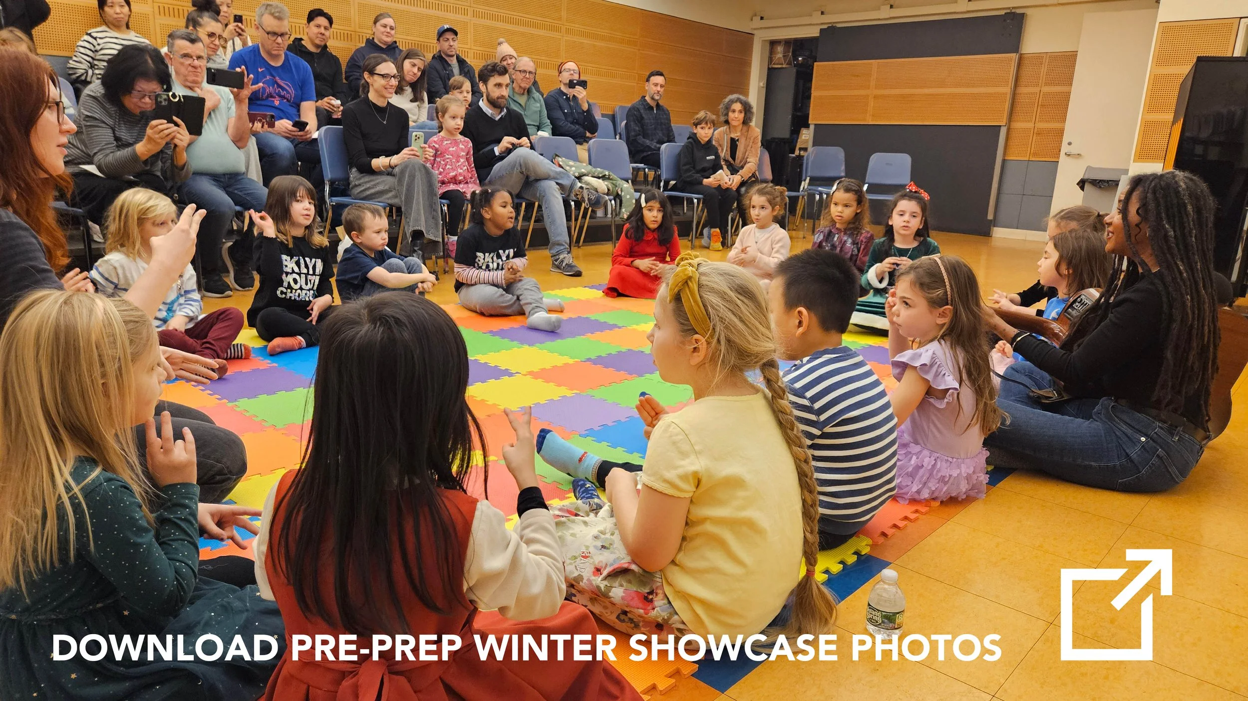 Children sitting on a colorful foam mat in a circle, with adults and parents sitting behind them, watching and recording a children's performance or event in a room with wooden panel walls.