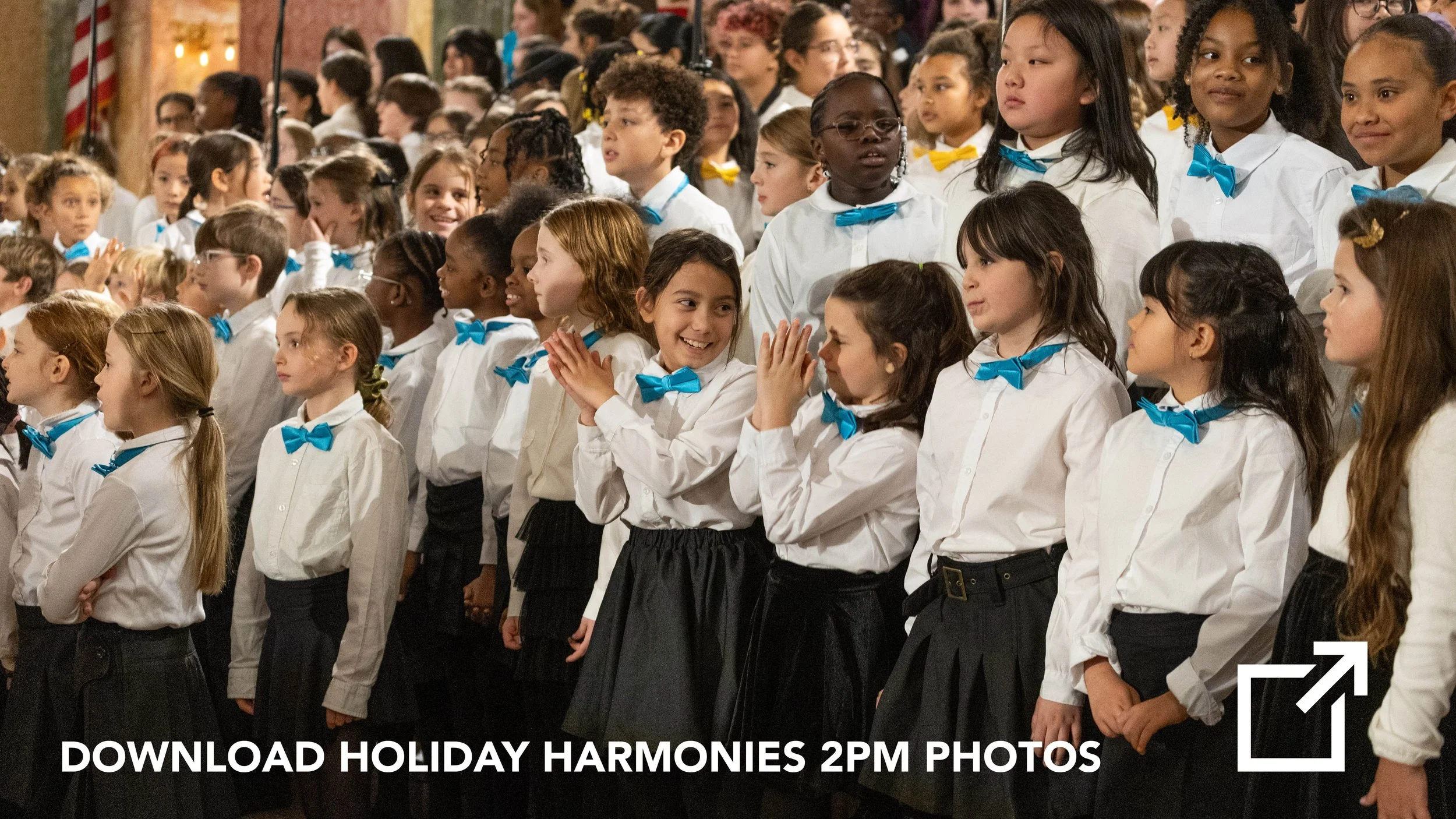 A large group of children dressed in white shirts and dark skirts or pants, some with blue bow ties, standing together and talking or smiling during a holiday event.