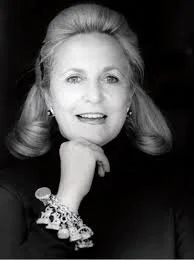 Black and white portrait of a woman with wavy hair, wearing a dark top and a large bracelet on her wrist, resting her chin on her hand.