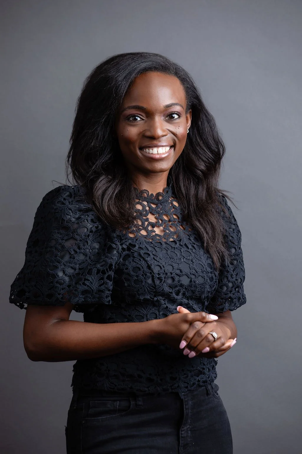 A woman smiling, wearing a black lace top with short sleeves, standing against a plain gray background.