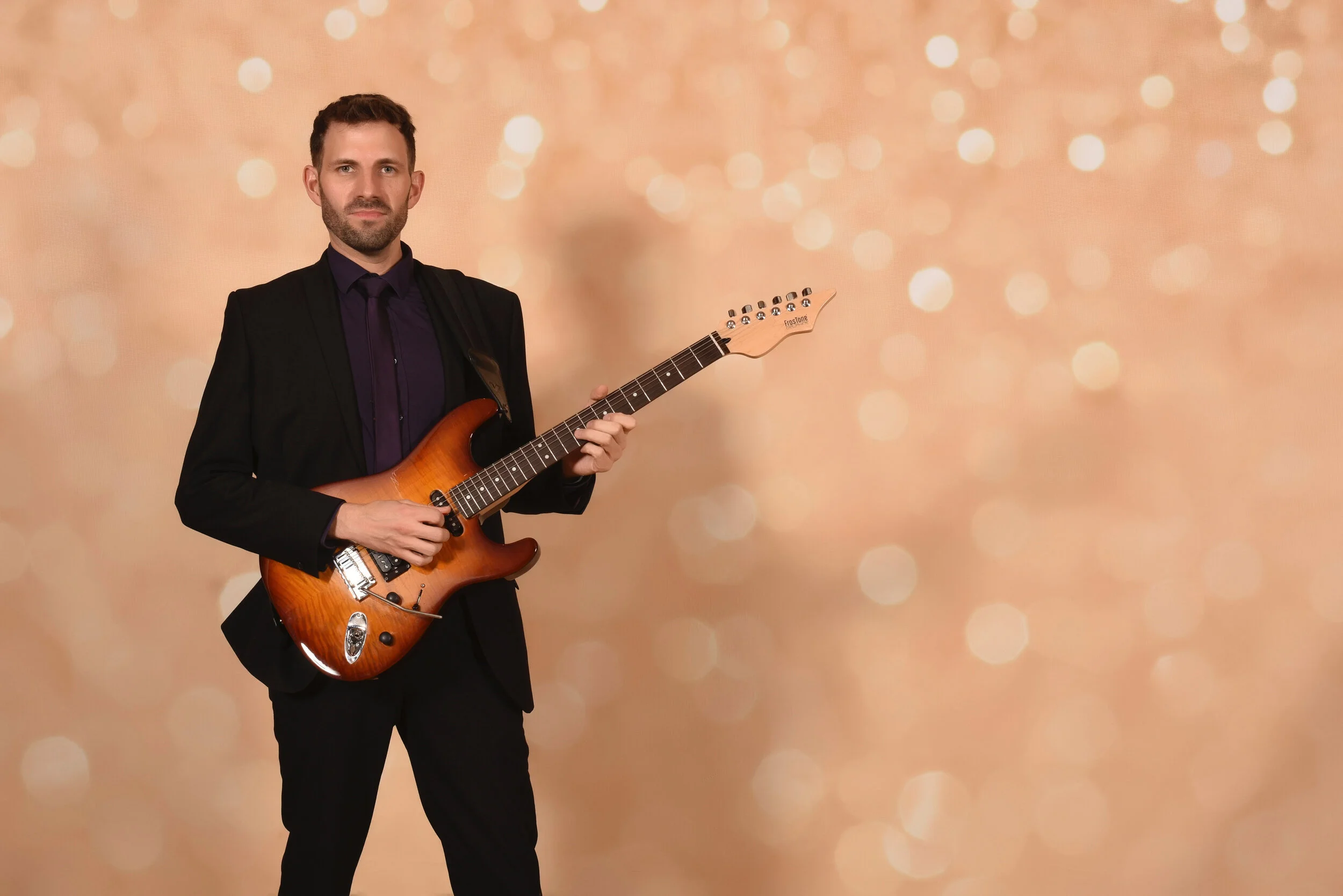 A man in a black suit holding an orange electric guitar standing against a bokeh background.