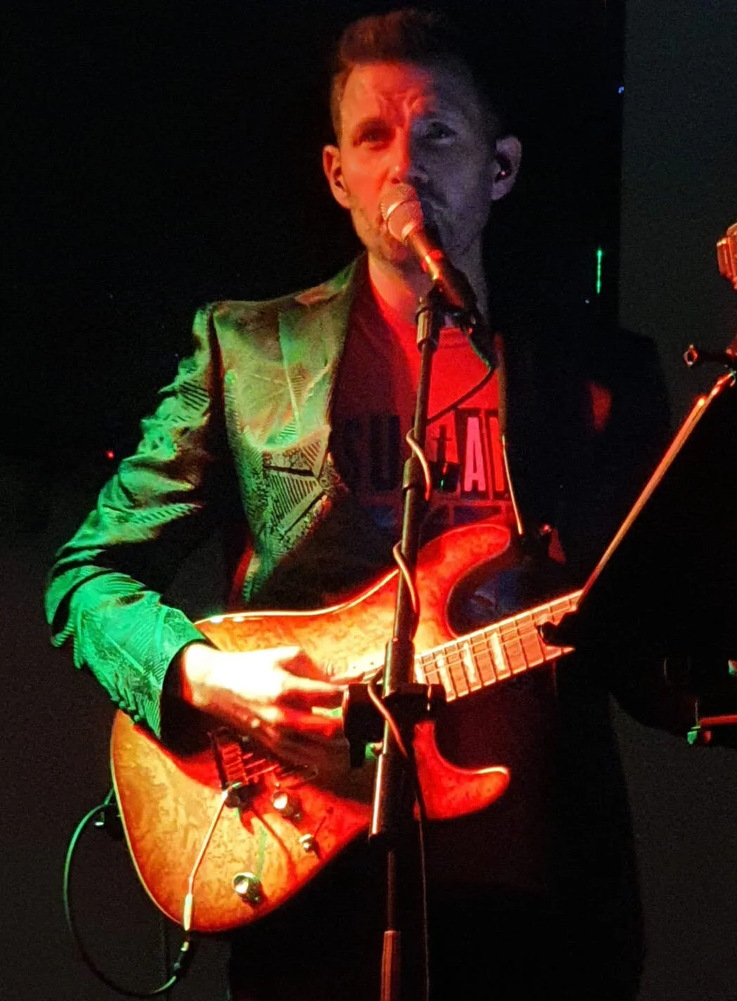A man playing an electric guitar and singing into a microphone on stage, with green and red stage lighting.