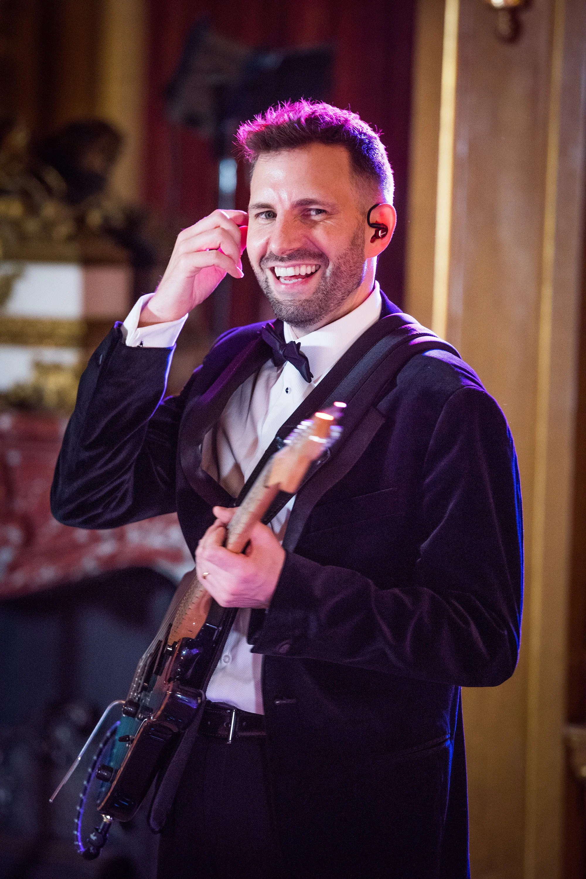 A man in a tuxedo and bow tie smiling while holding a guitar, adjusting an earpiece in his ear with one hand, in an elegant room with ornate decor.
