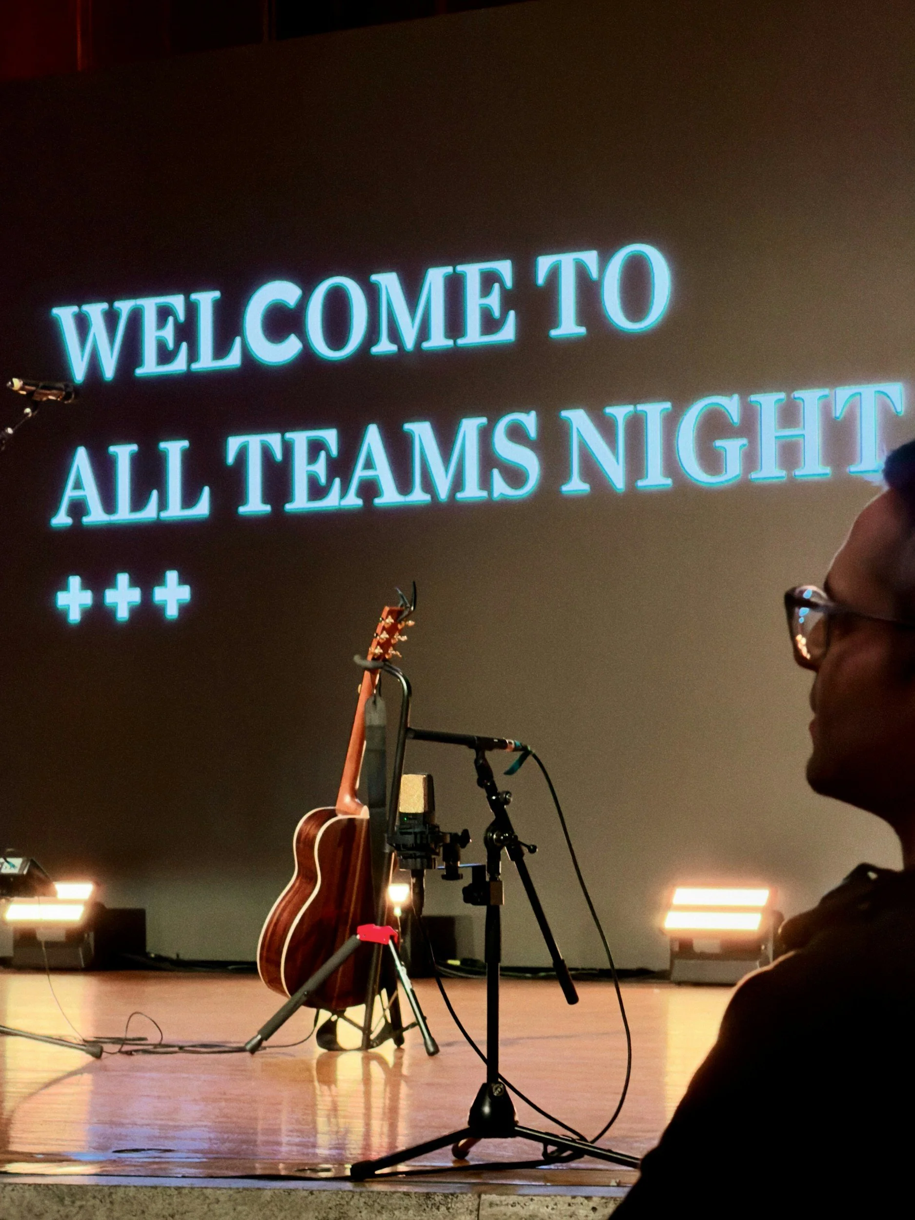 Stage with a guitar on a stand, microphone, and bright stage lights, with a large screen in the background displaying a message that says "Welcome to All Teams Night" with three plus signs underneath.