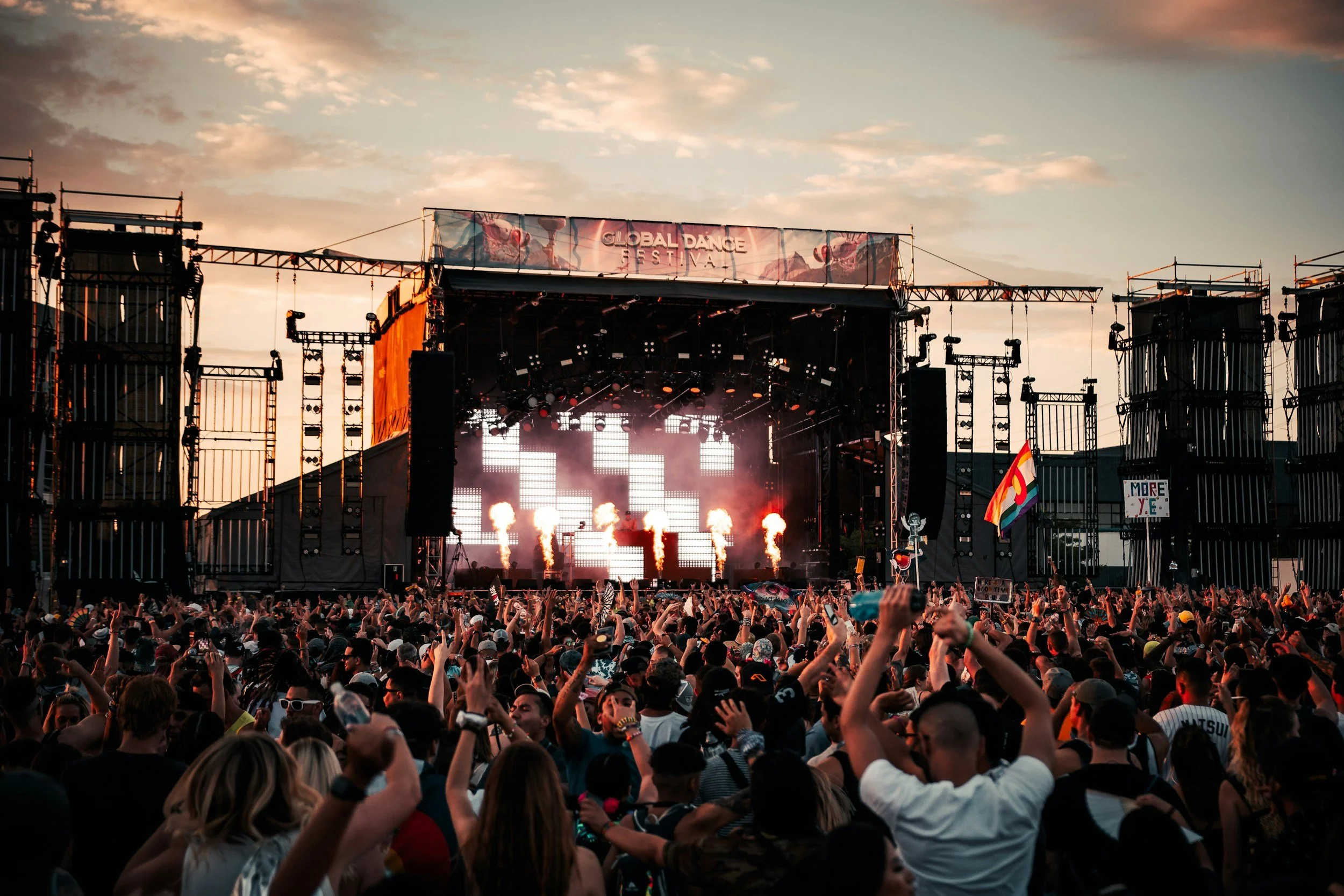 Large crowd attending an outdoor music festival at sunset, with a stage featuring bright lights, pyrotechnics, and a 'Global Dance Festival' banner.