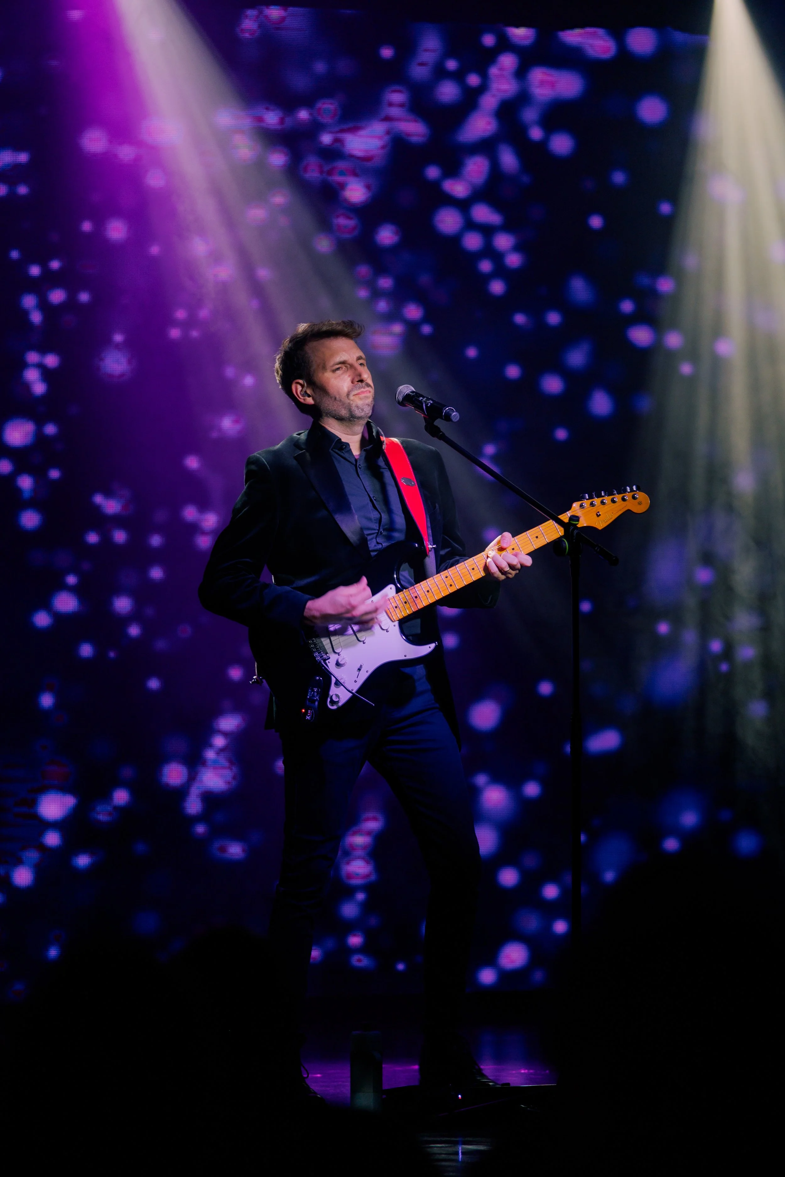 Ian Michael Frost performing on stage, light rays, blue and purple, guitarist standing holding a Fender Stratocaster guitar, purple spots background,