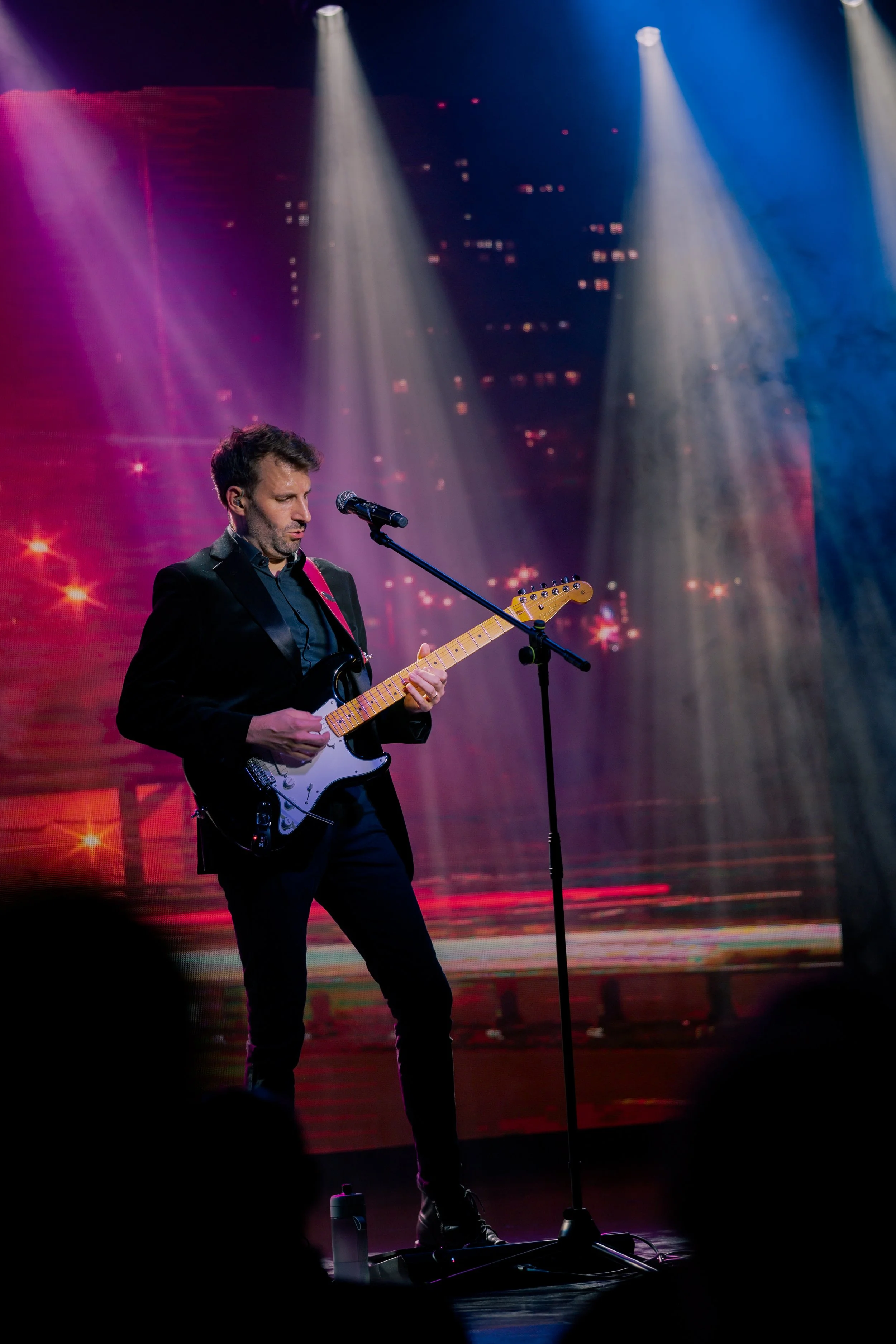 Male musician playing electric guitar on stage with colorful lighting and cityscape projection in background.