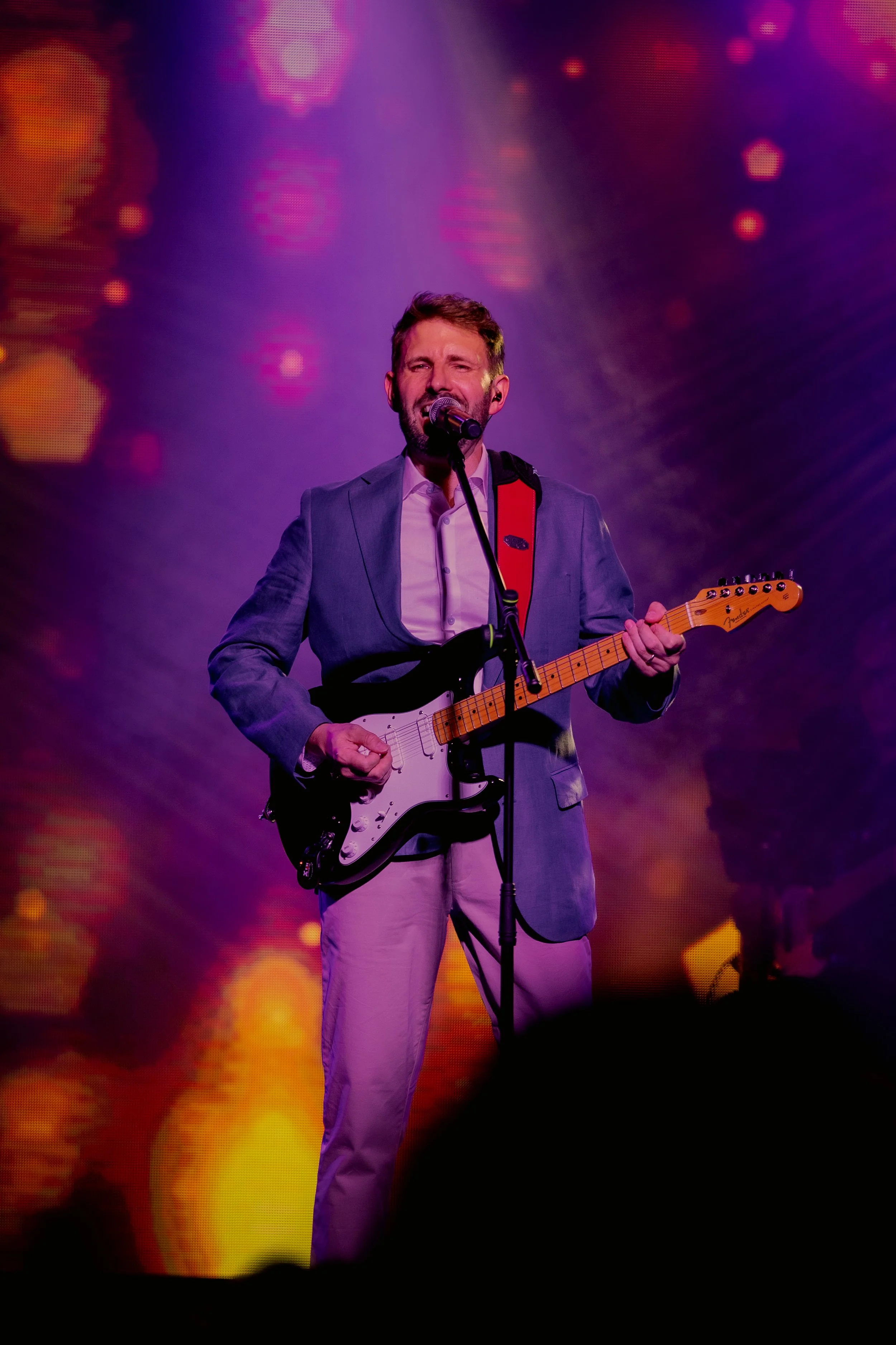Ian Michael Frost performing on stage, spotlight, purple, gold, orange, guitarist standing holding a Fender Stratocaster guitar, singing into a microphone