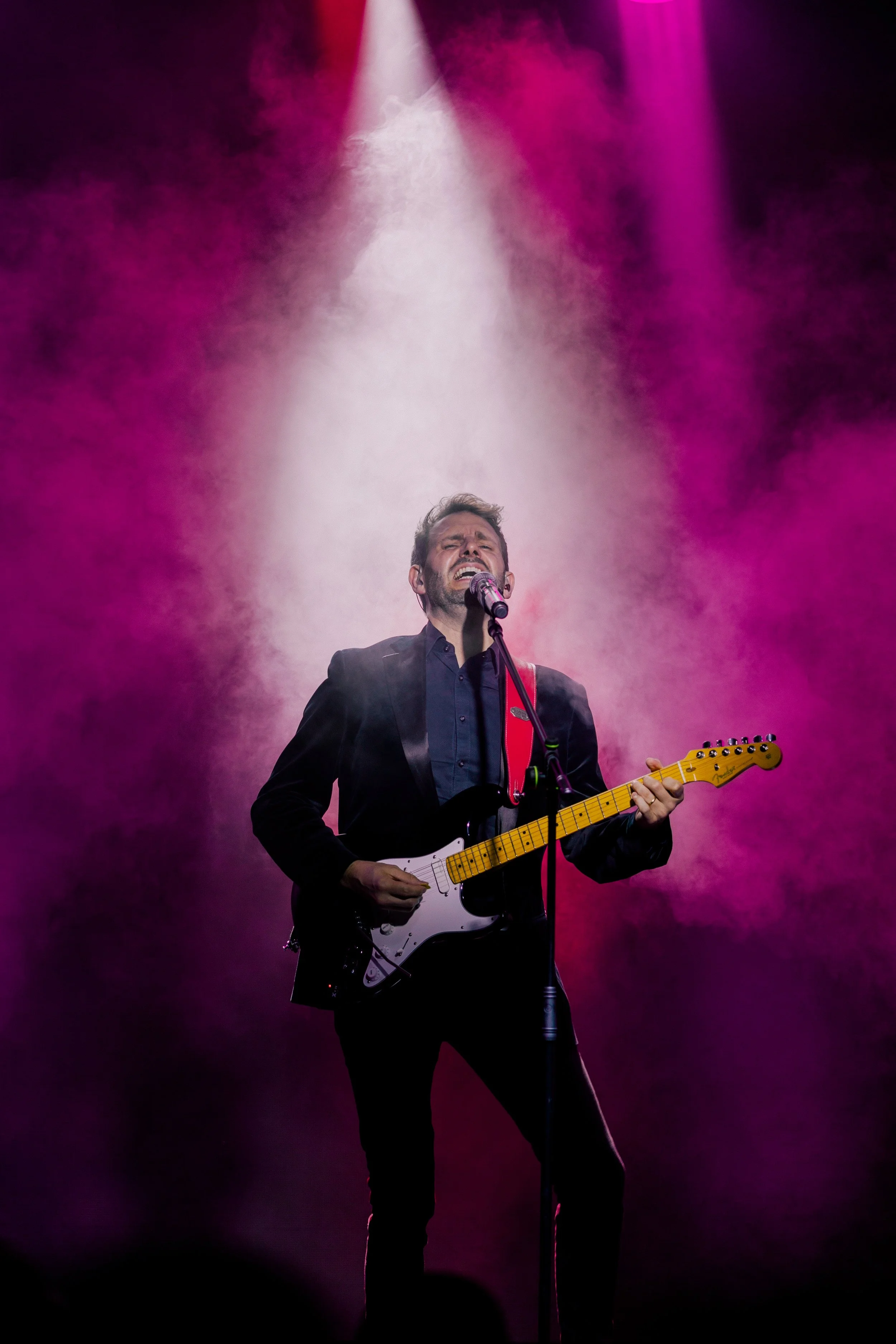 Ian Michael Frost performing on stage, spotlight, pink and purple, guitarist standing holding a Fender Stratocaster guitar, singing into a microphone