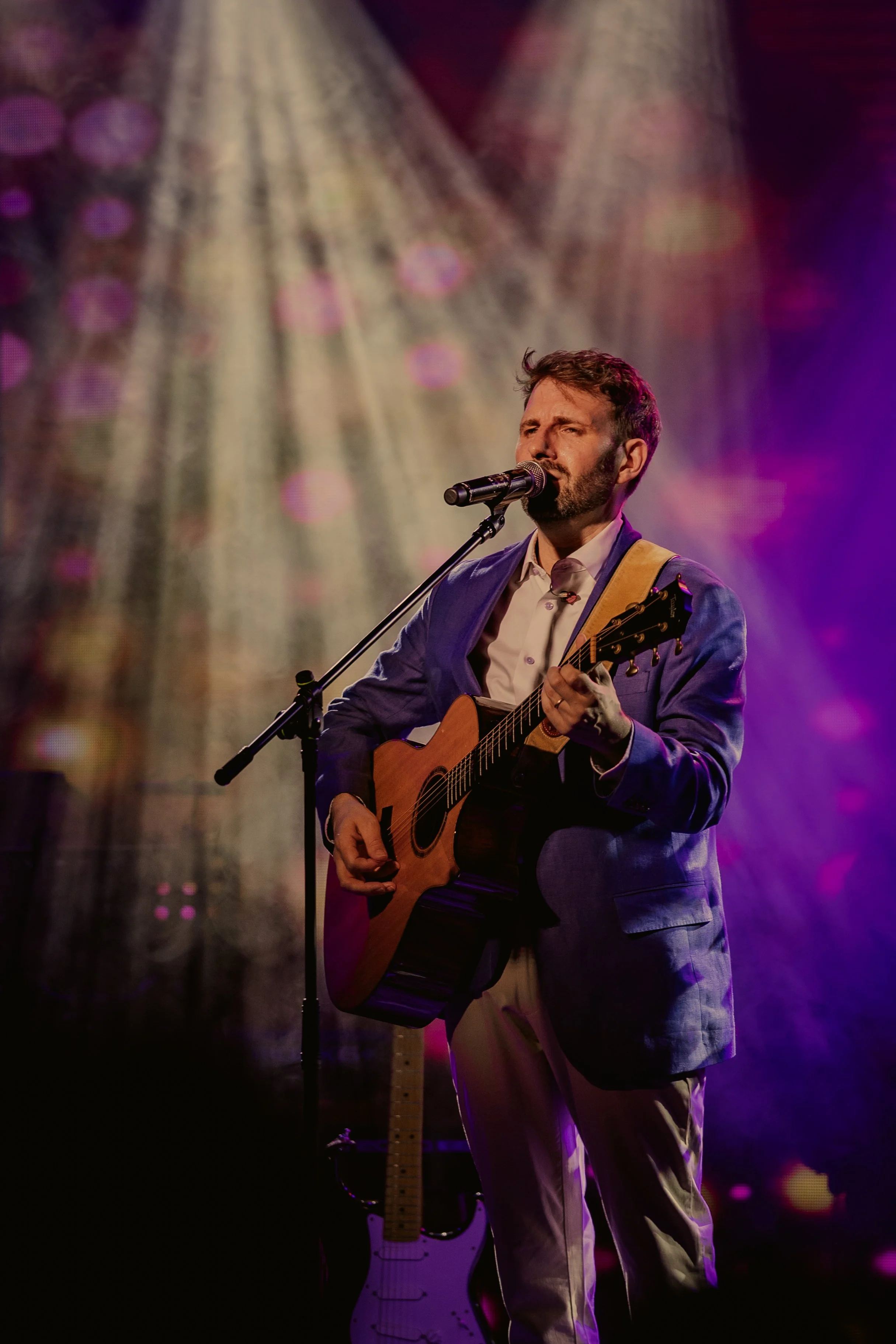 Ian Michael Frost performing on stage, spotlight, white, pink and purple light, guitarist standing holding an acoustic guitar, singing into a microphone, three-quarter view,