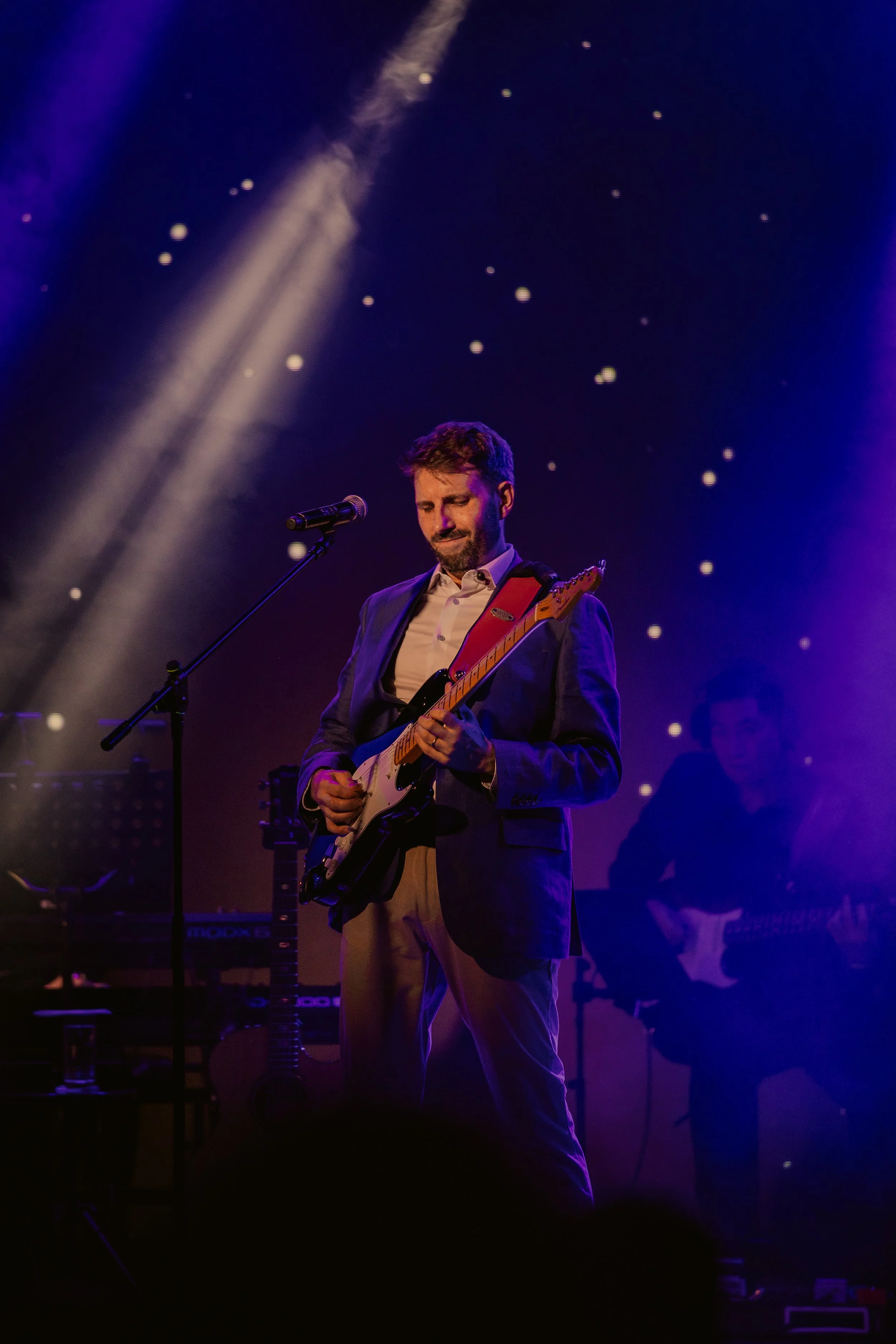 Ian Michael Frost performing on stage, light rays, blue and purple, guitarist standing holding a Fender Stratocaster guitar, starlight background