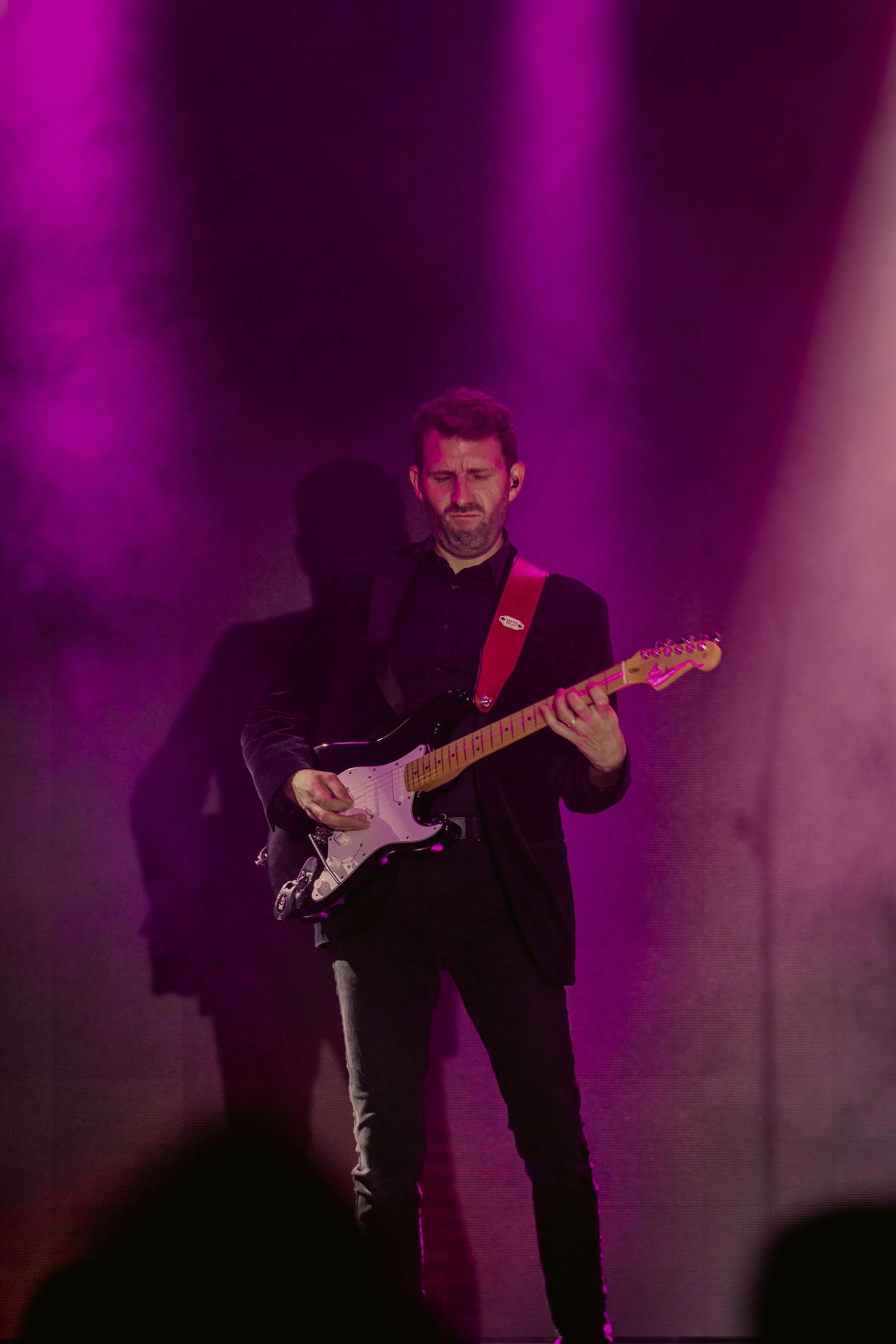 Ian Michael Frost performing on stage, dark suit, guitarist standing holding a Fender Stratocaster guitar, looking down, front view, light rays, pink light,