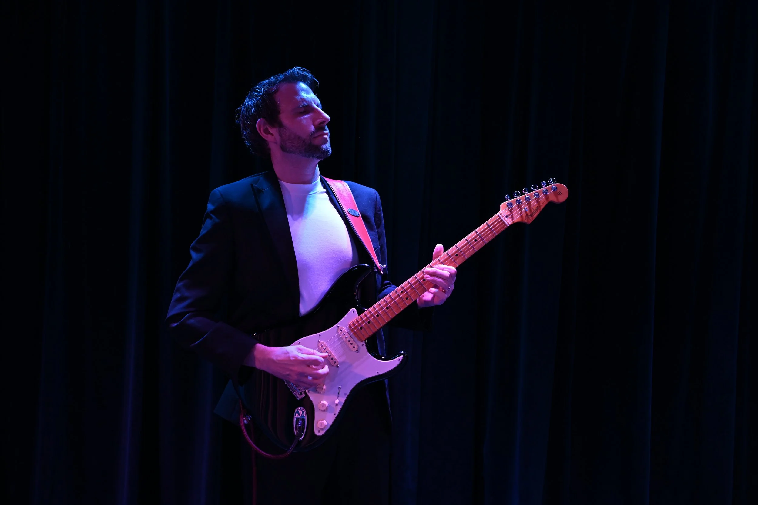 A man with dark hair and a beard playing an electric guitar on stage with dark curtains behind him, illuminated by purple and blue stage lights.