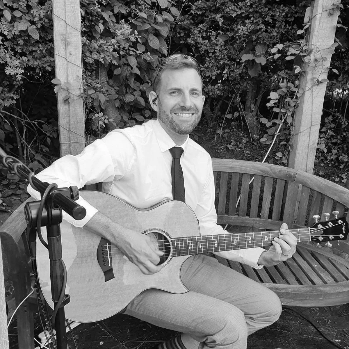A man with a beard sitting on a wooden park bench, smiling, playing an acoustic guitar. He is wearing a white shirt, tie, and light-colored pants, with sunglasses on his head. The background has dense foliage and trees.
