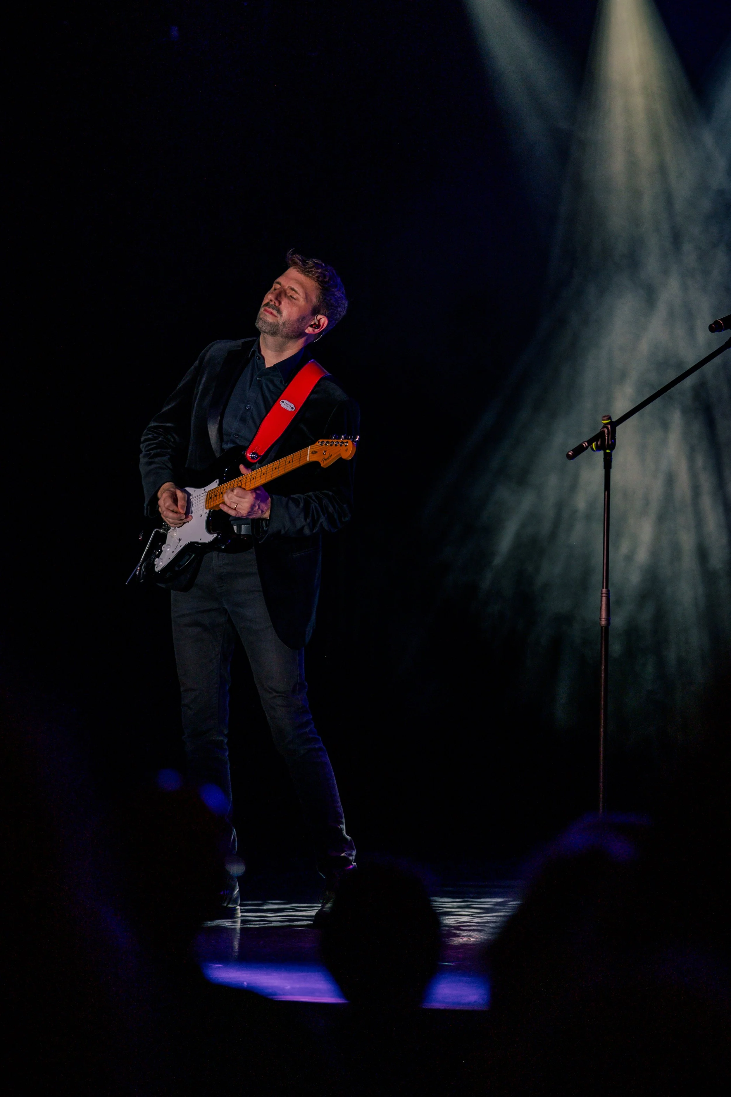 Ian Michael Frost performing on stage, light rays, white lights on a dark stage, guitarist standing holding a Fender Stratocaster guitar, looking up, eyes closed,