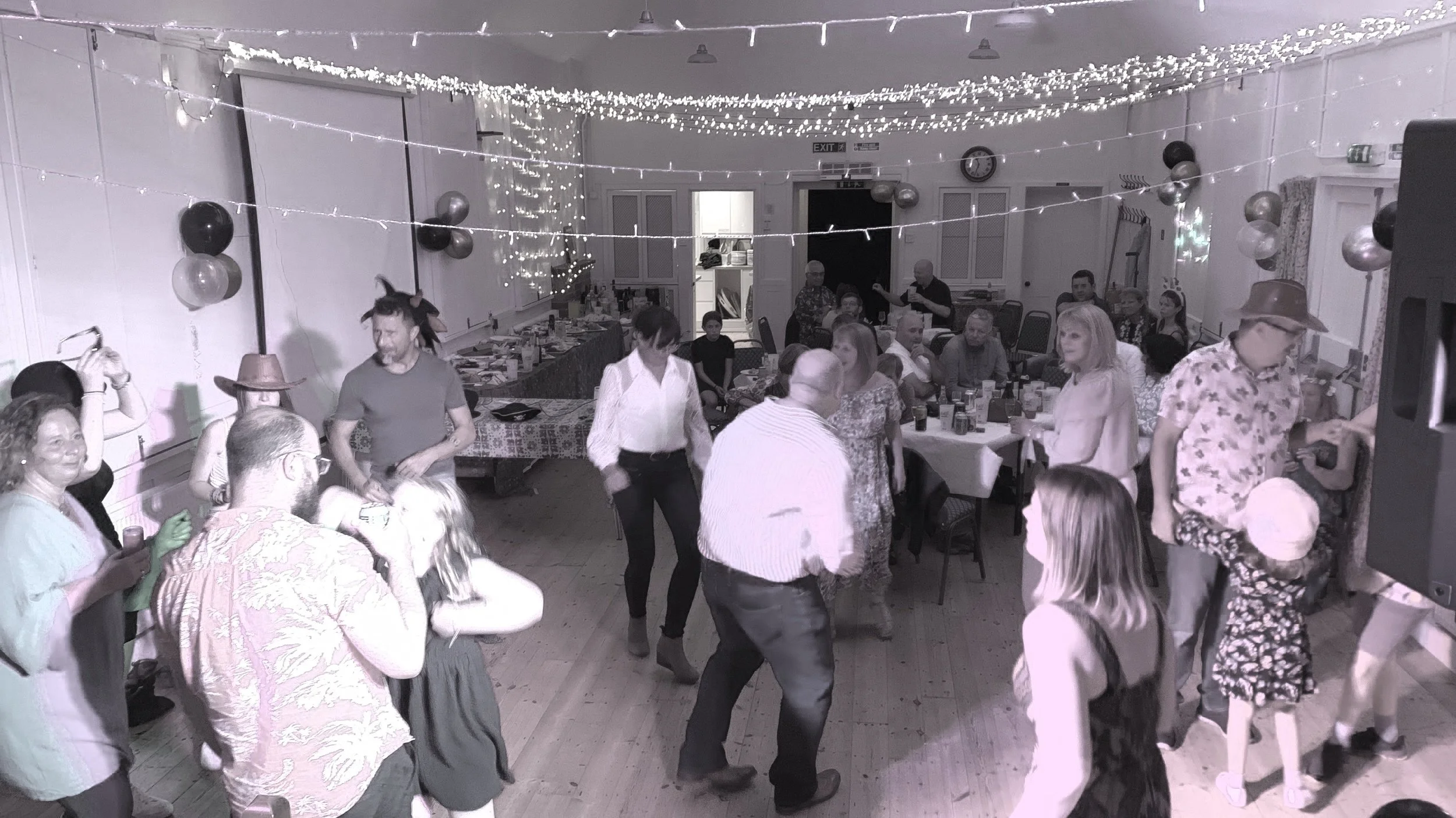People dancing and socializing at a celebration or party in a decorated hall with string lights and balloons.