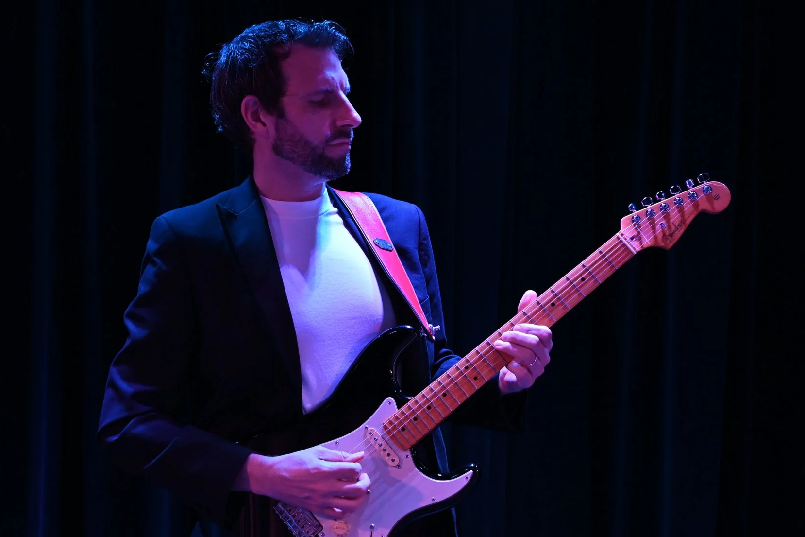 A man playing an electric guitar on stage with dark curtains in the background.
