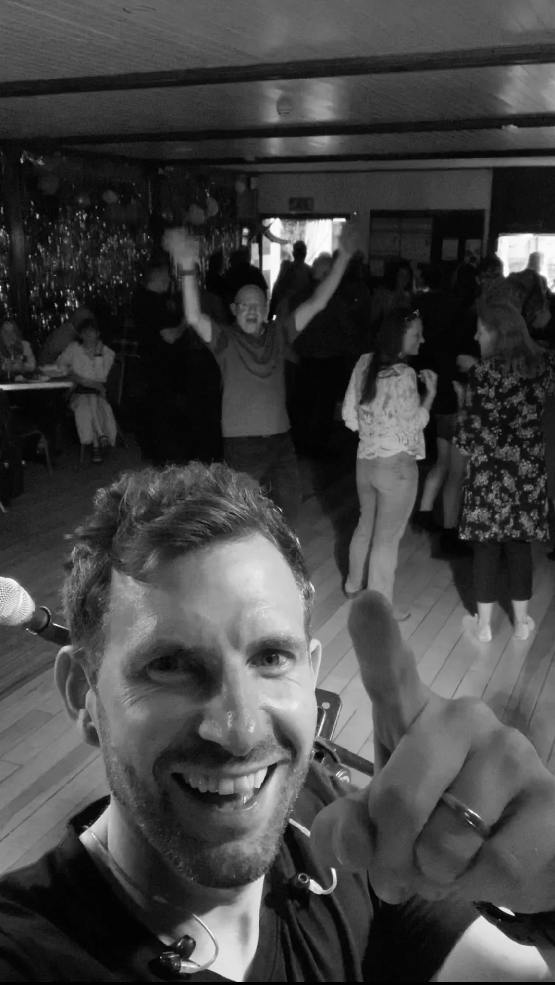 A man smiling and pointing at the camera during a lively celebration or party, with people dancing and socializing in the background.