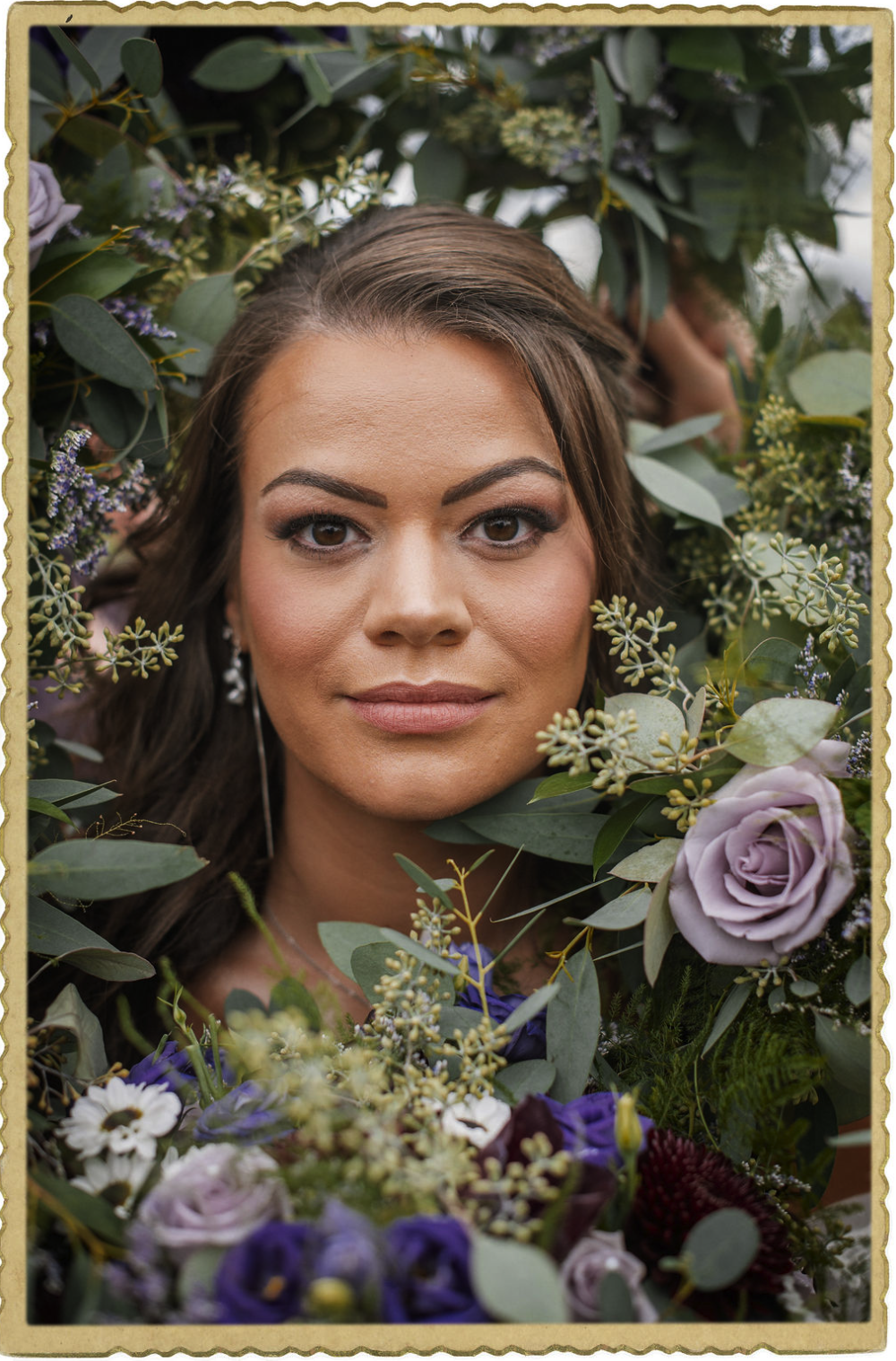 A woman with light skin, brown hair, and makeup, posing among green leaves, purple, white, and burgundy flowers, with a soft-focus background.