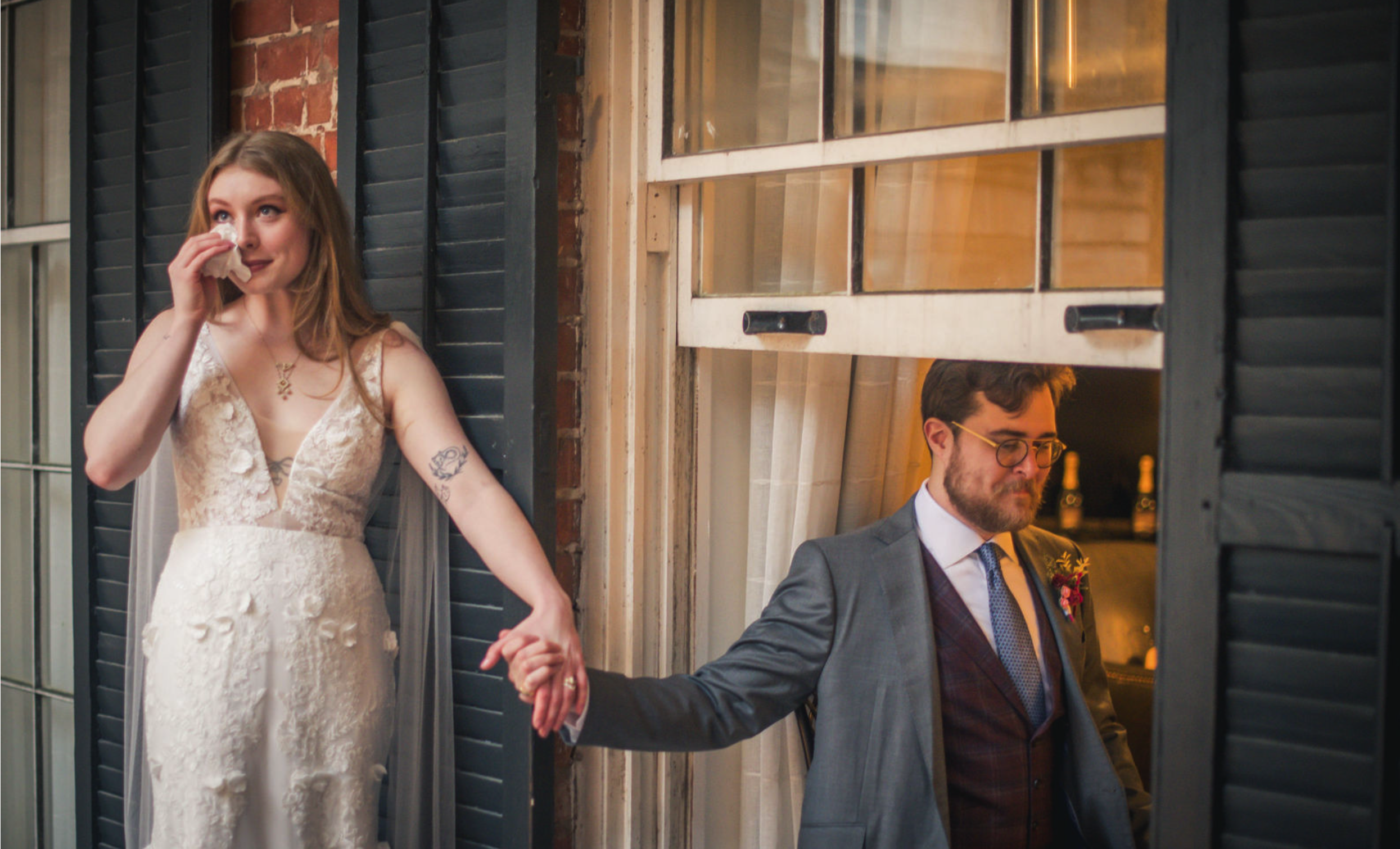 A woman in a wedding dress holding hands with a man in a gray suit who is looking down, seen through an open window with black shutters.