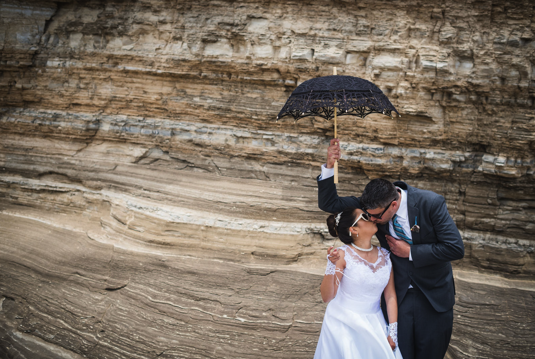 Couple in wedding attire sharing a kiss under a black lace umbrella against a rocky landscape backdrop.