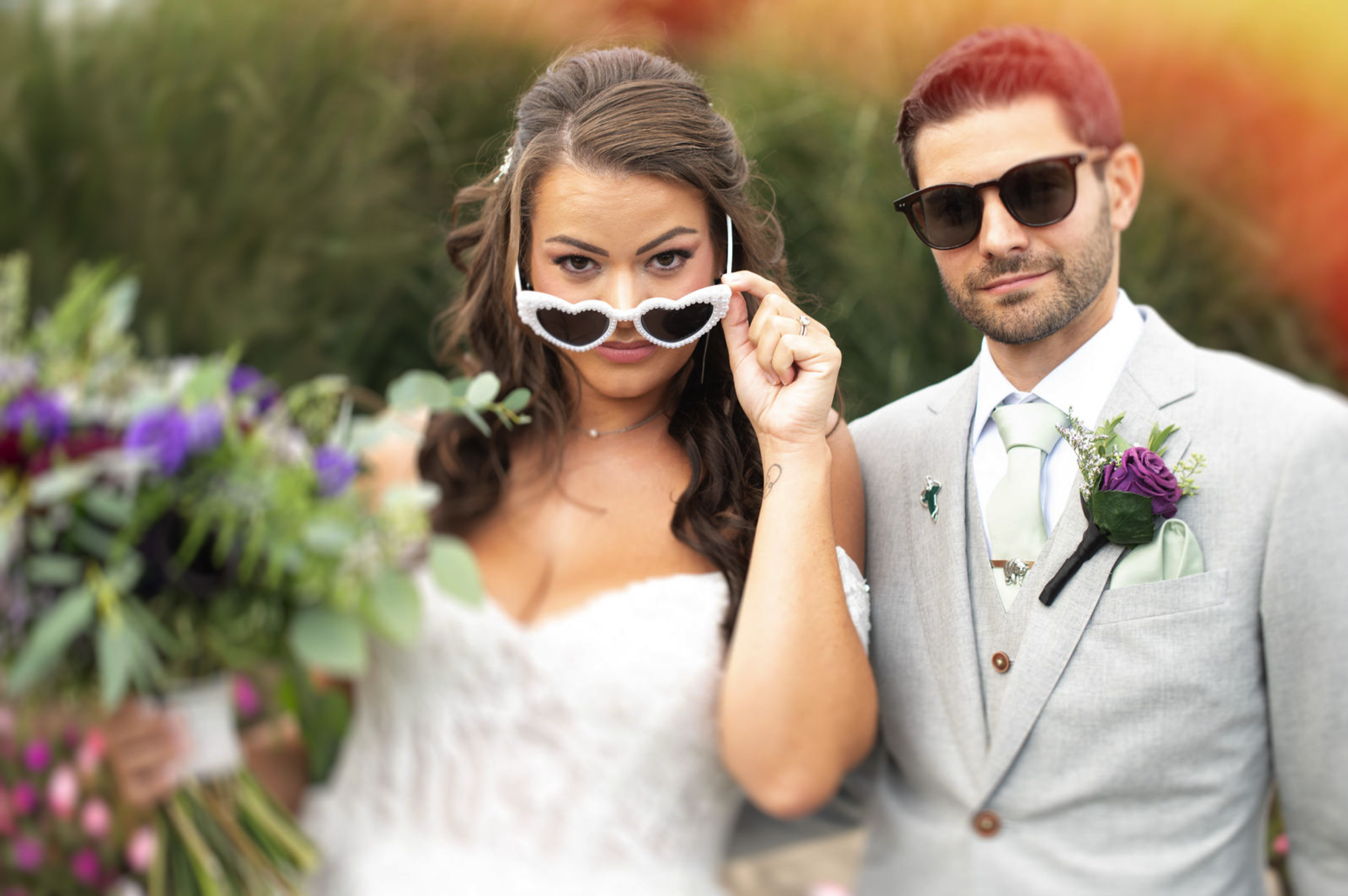 A bride and groom wearing sunglasses outdoors, with the bride holding a bouquet and adjusting her heart-shaped sunglasses, and the groom dressed in a light gray suit with a purple boutonniere.