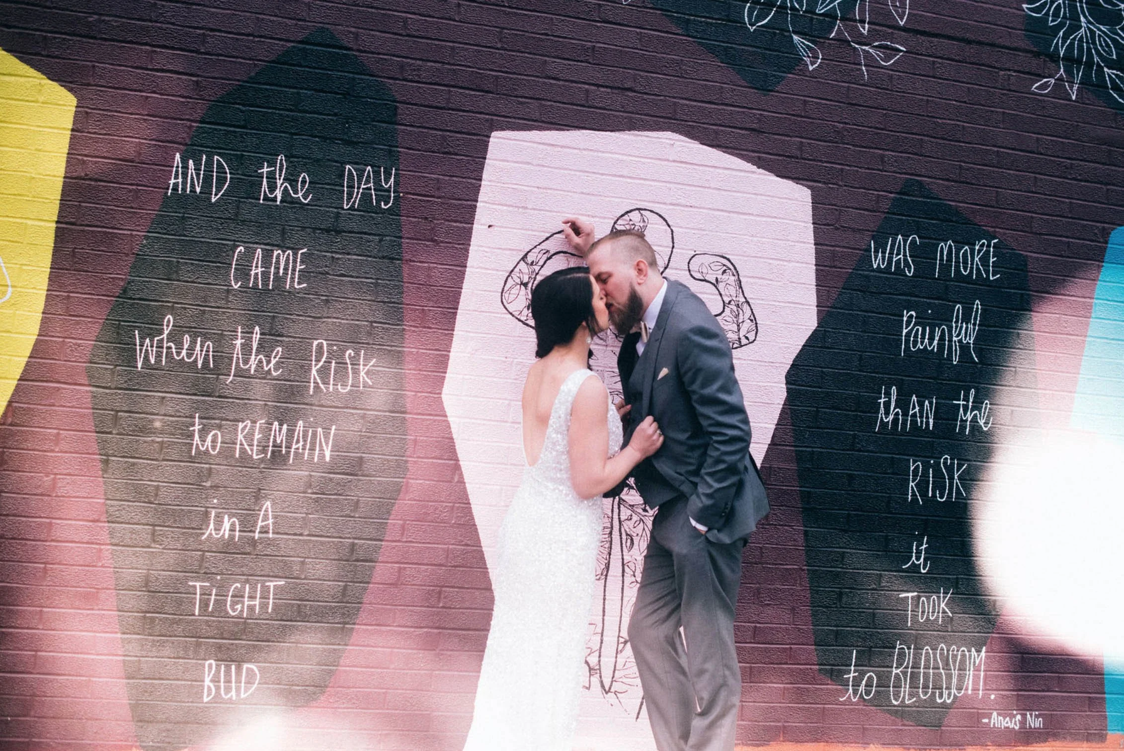 A bride and groom kissing in front of a colorful mural on a brick wall, with handwritten-style quotes on either side.