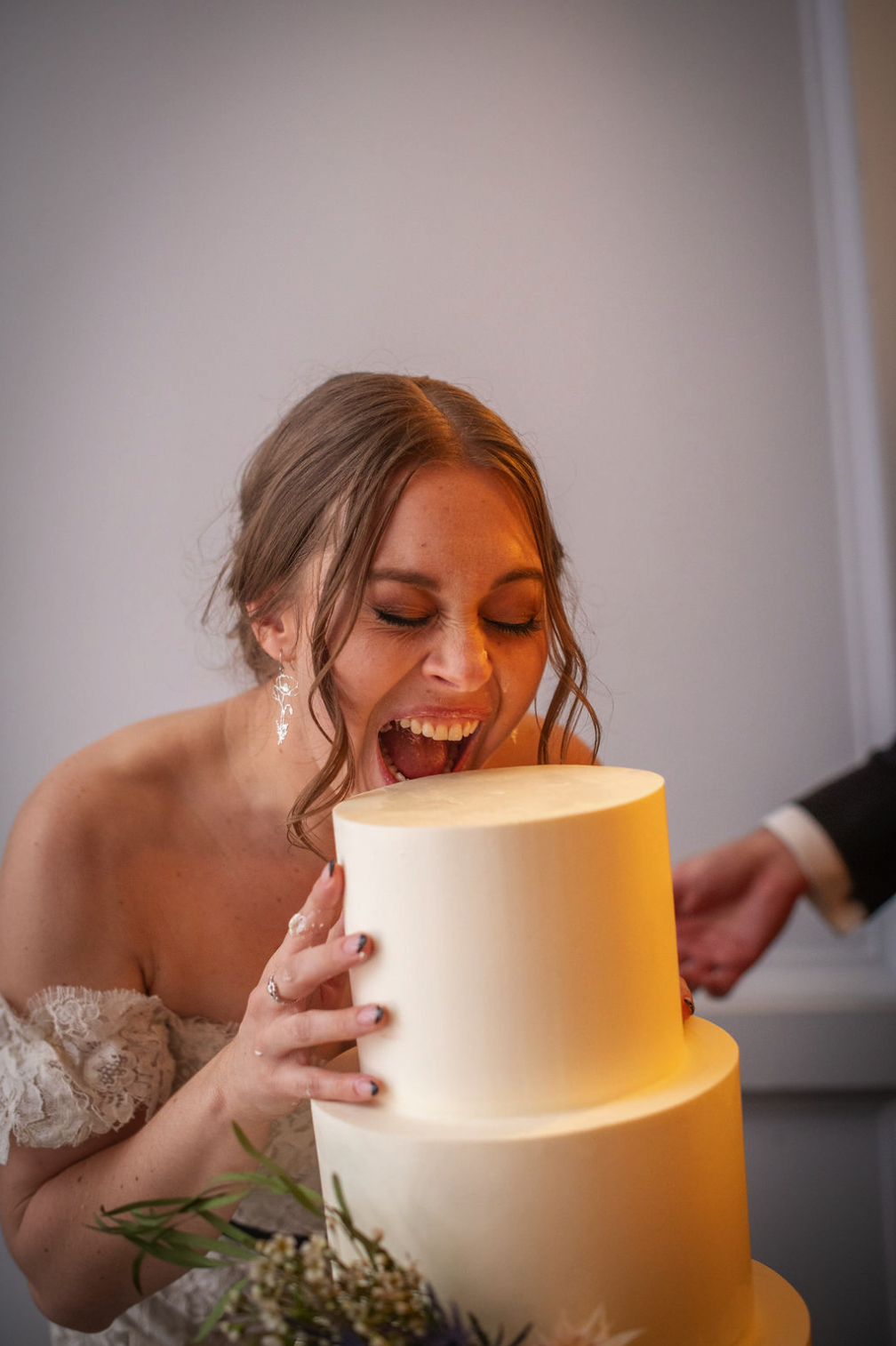 A woman in a wedding dress is pretending to eat a large, tiered wedding cake at her wedding reception.